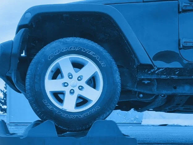 Close-up of a black vehicle's front tire mounted on a wheel, resting on a wheel chock, with a blue filter applied.