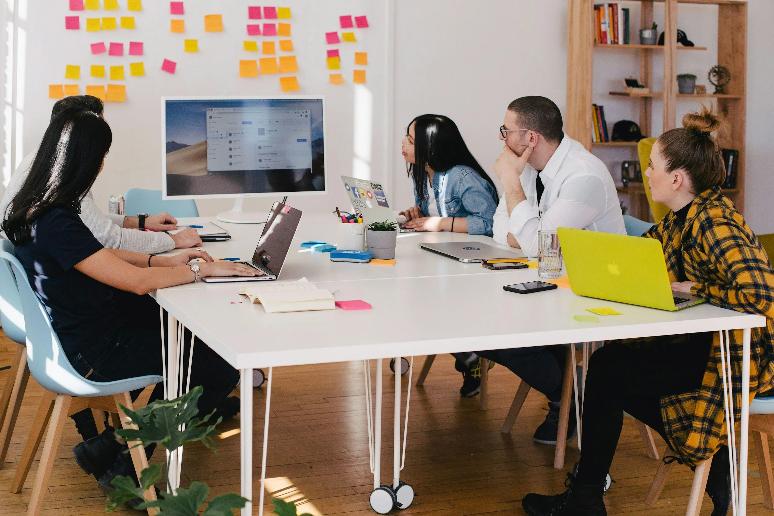 A group of five people sitting around a white table in a modern office, working on laptops and engaged in a meeting. There is a large monitor on the table and sticky notes on the wall behind them. The office has wooden floors, a bookshelf, and various office supplies.