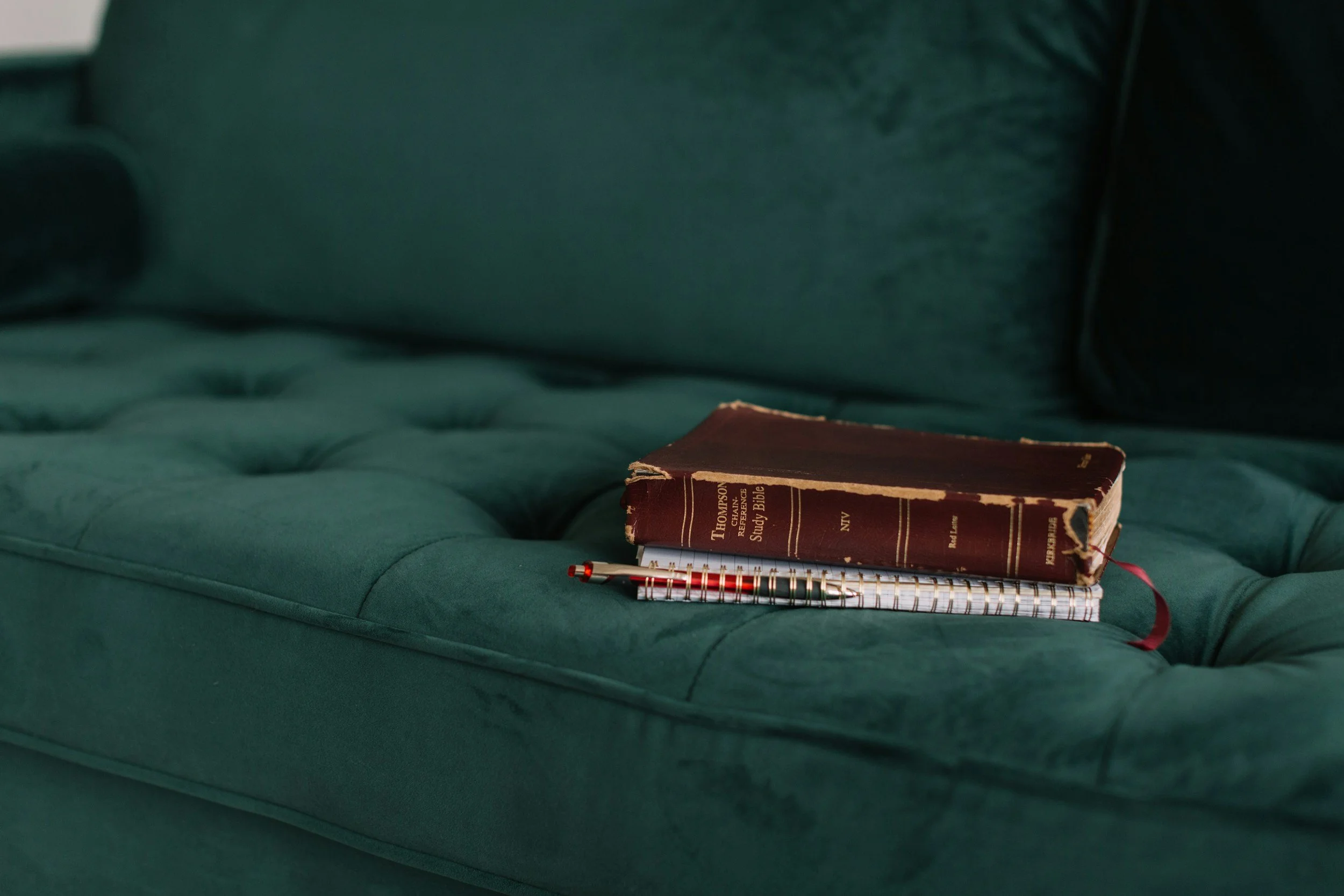 A worn, red hardcover Bible titled 'Thompson Chain Reference Study Bible' resting on a green tufted sofa, with a white spiral notebook and a red pen placed on top.