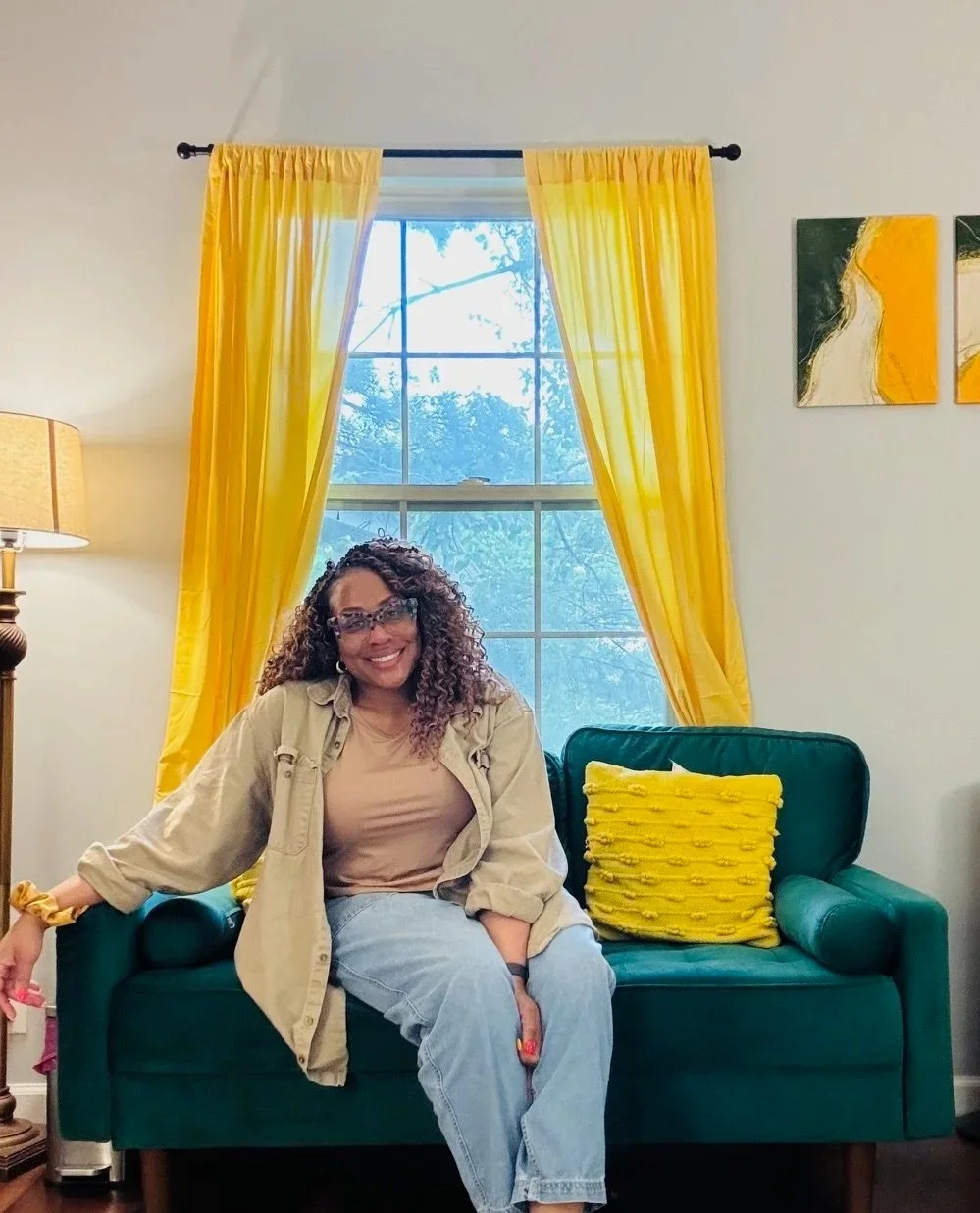 A woman with curly hair and glasses, smiling, sitting on a green sofa with a yellow pillow, in front of yellow curtains and a large window with tree views in a well-lit room.