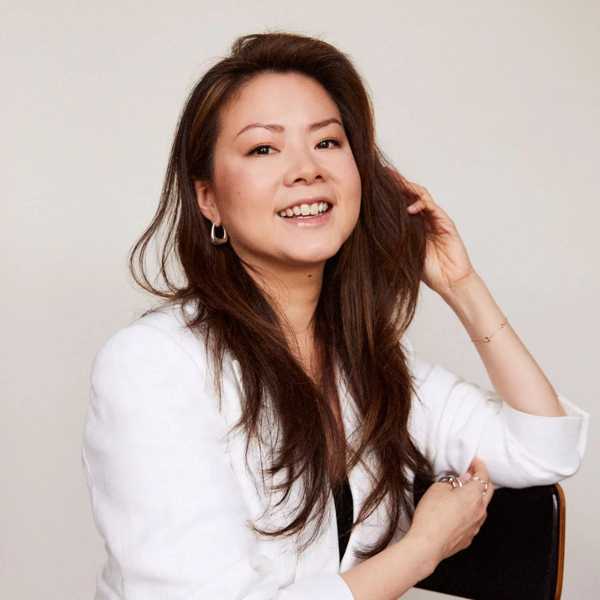 Portrait of a smiling woman with long brown hair, wearing a white blazer, earrings, and bracelets, sitting against a plain light background.