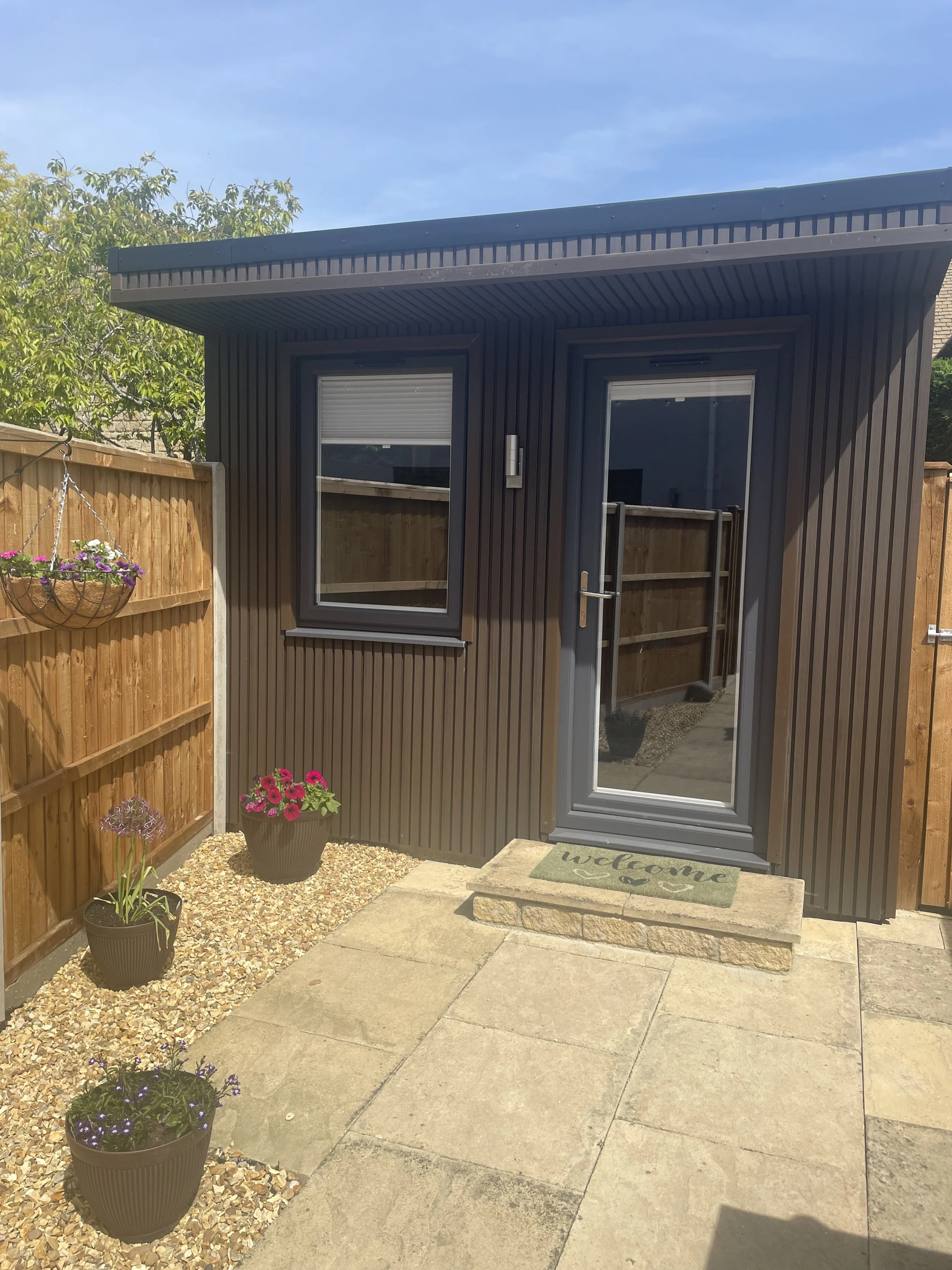 A small backyard shed with a black metal exterior and a glass door. The shed is situated next to a wooden fence with hanging flower pots. The ground is a mix of gravel and concrete, with potted flowers and a welcoming doormat.