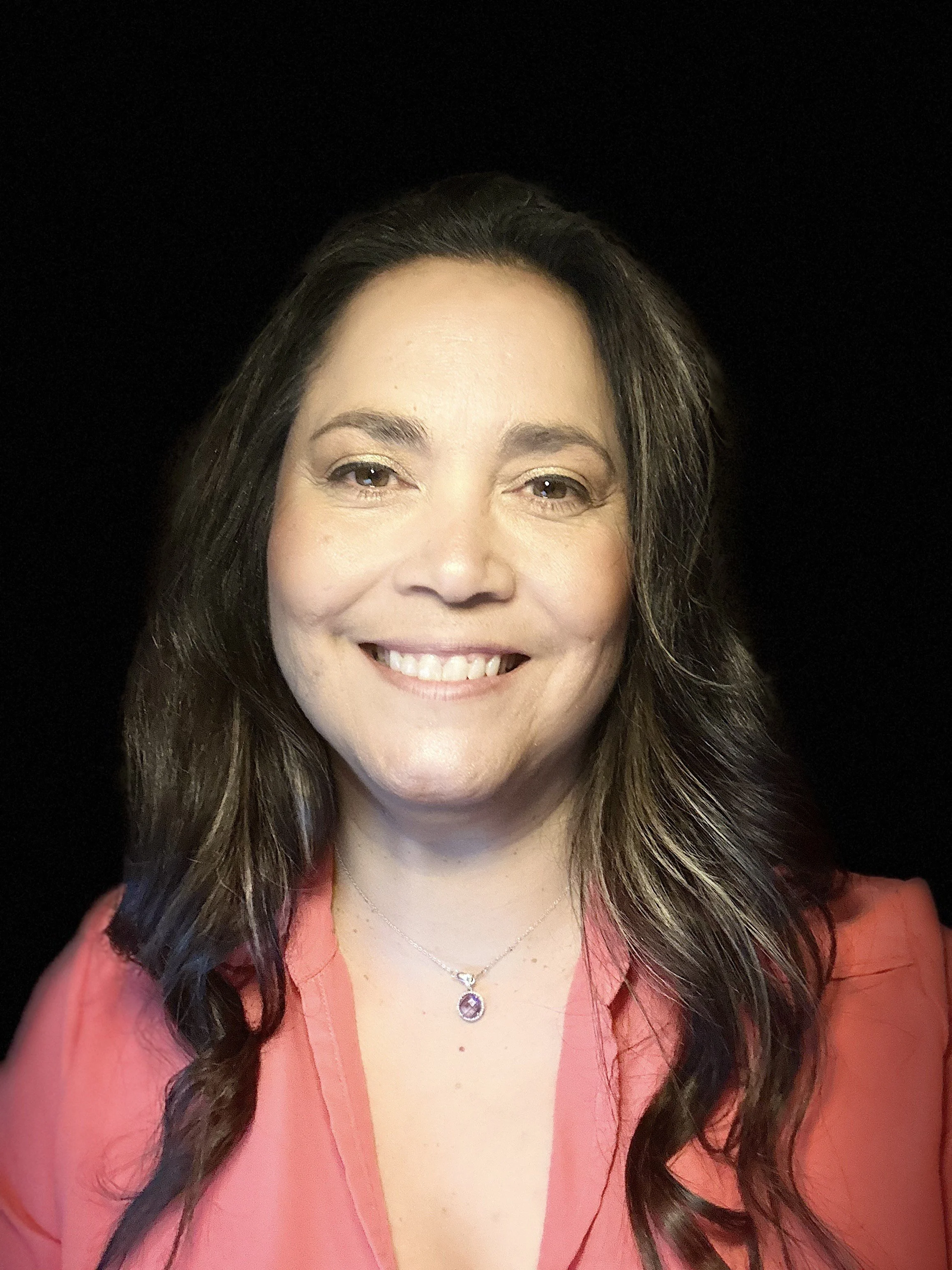 Smiling woman with long hair wearing a coral top and necklace with a purple pendant.