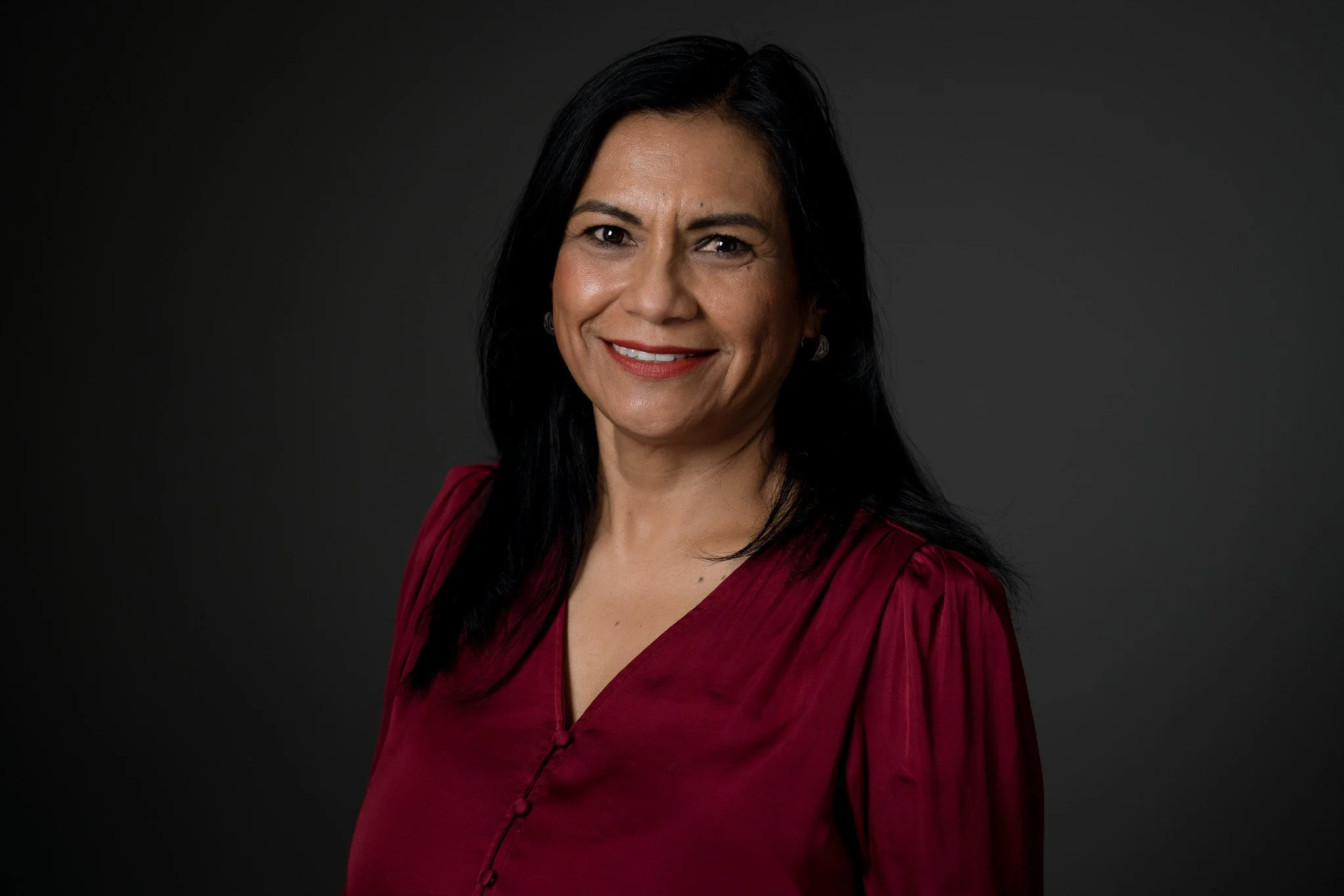 Portrait of a woman with dark hair wearing a maroon blouse, smiling against a dark background.