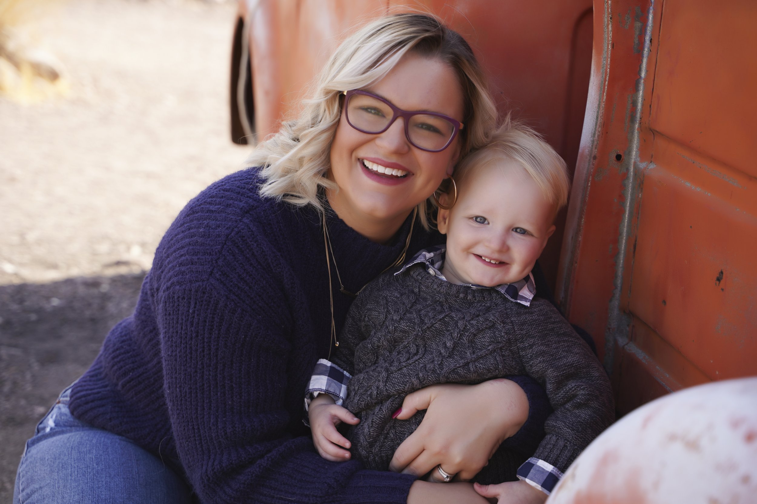 A woman and a young boy sitting outside leaning against a red wall, both smiling.