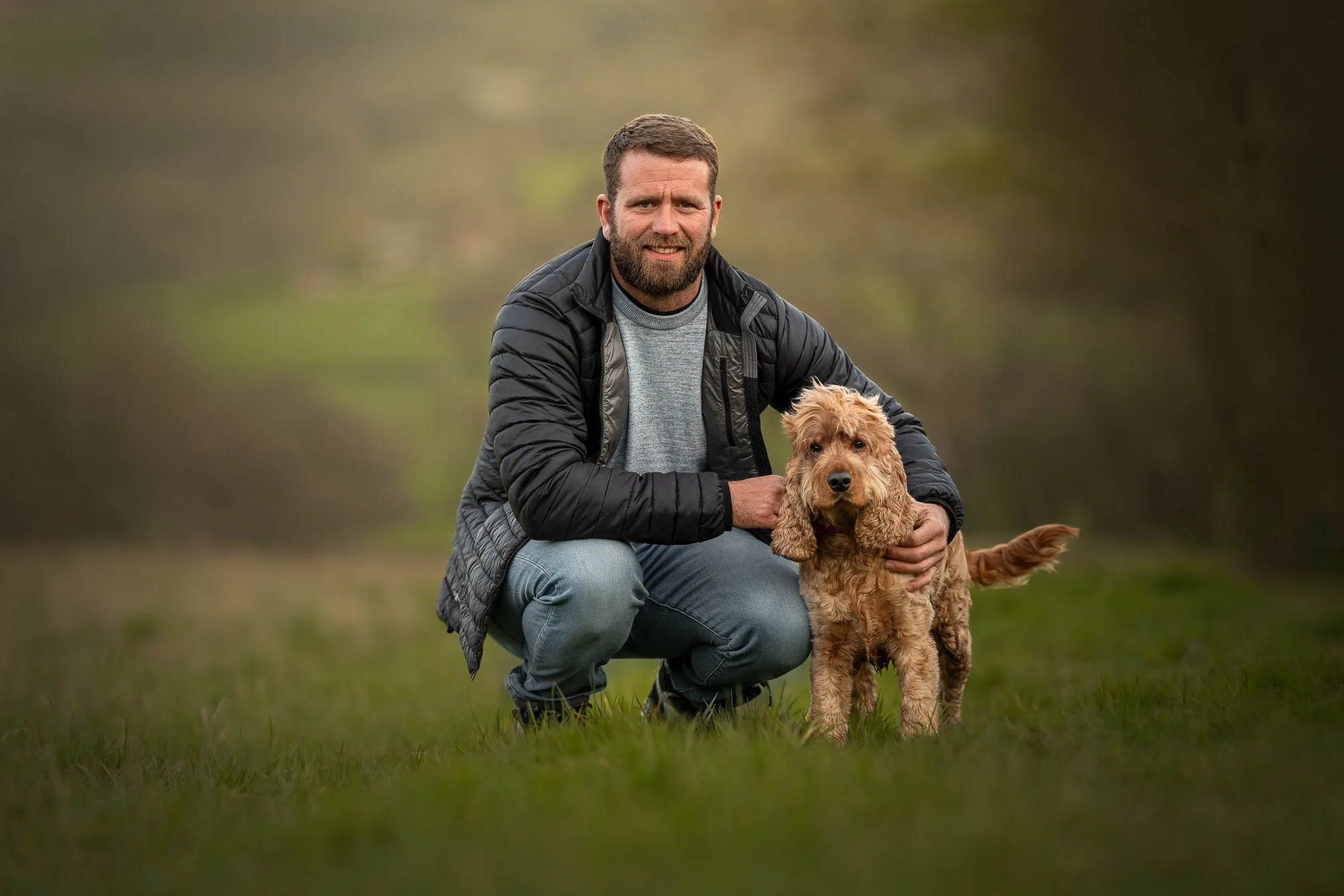 A man crouching outdoors with a tan, curly-haired dog during daytime with a blurred natural background.