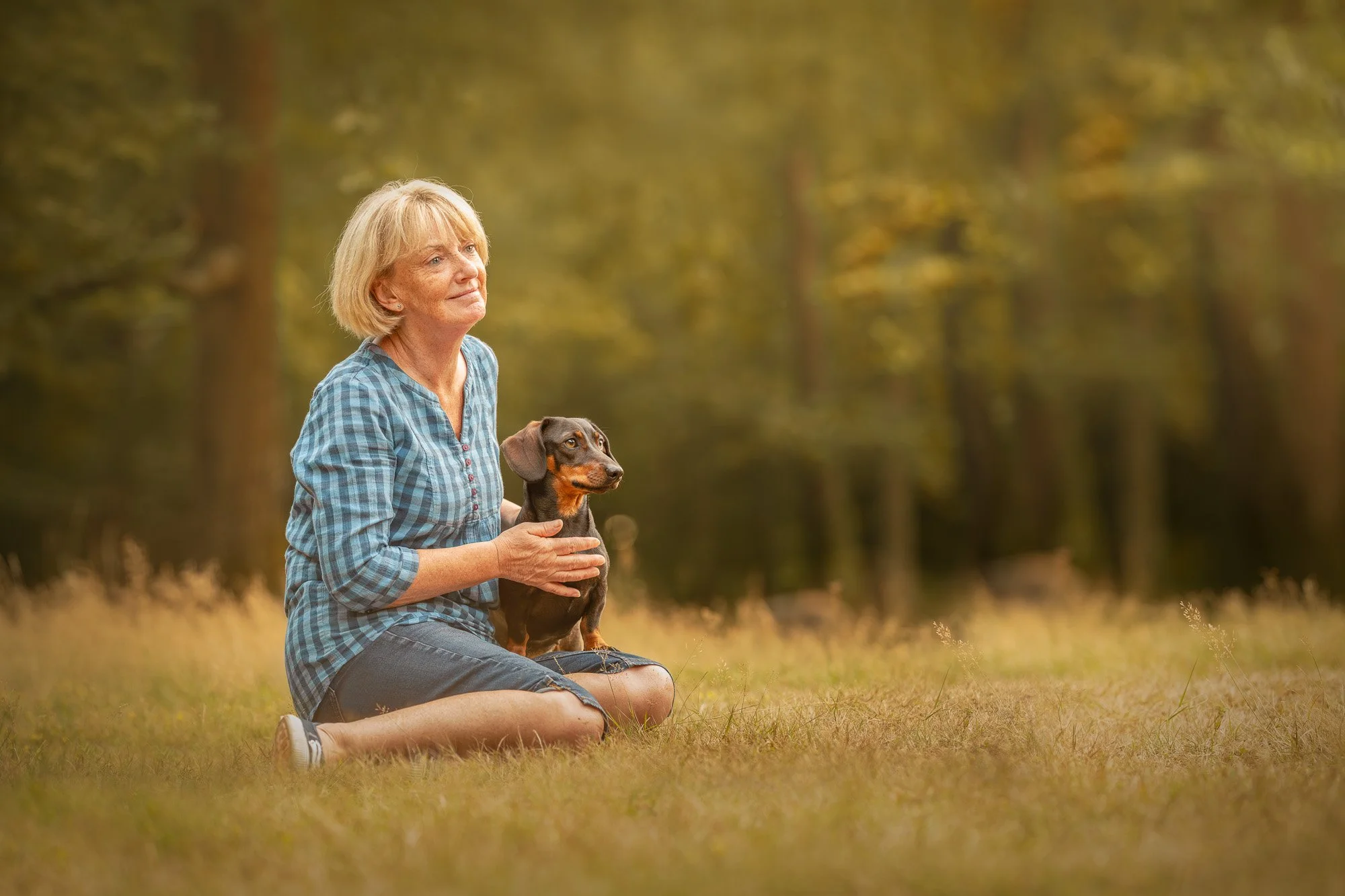 An older woman sitting on the grass in a forest, holding a small dachshund dog, both looking peacefully into the distance.