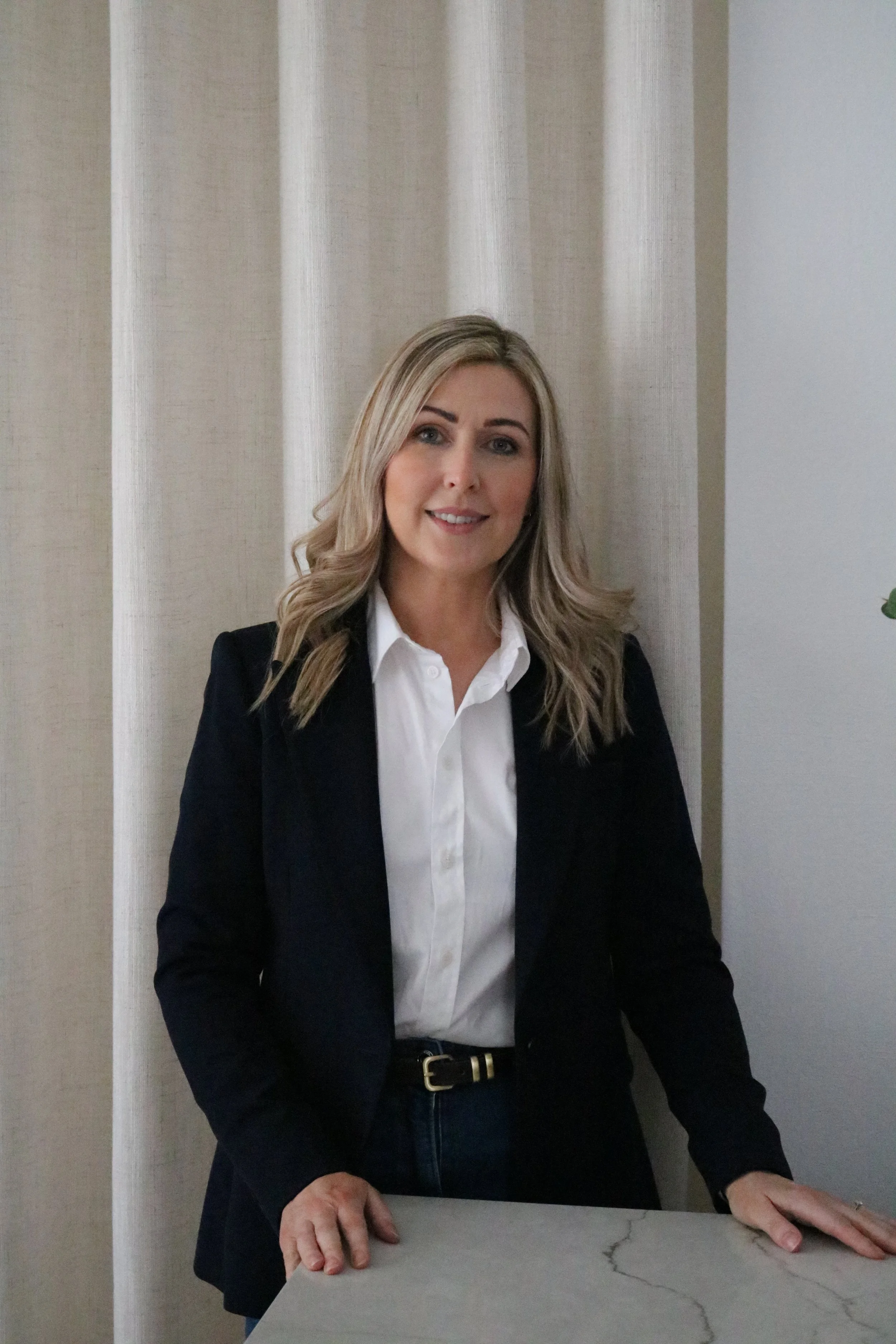 A woman with blonde hair wearing a navy blazer over a white shirt standing behind a white marble table.