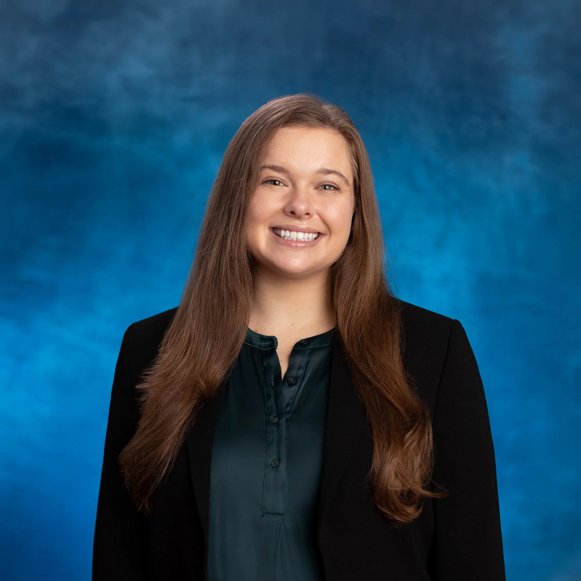 Close-up portrait of a smiling woman with long brown hair, wearing a black blazer and dark green blouse, against a blue background.