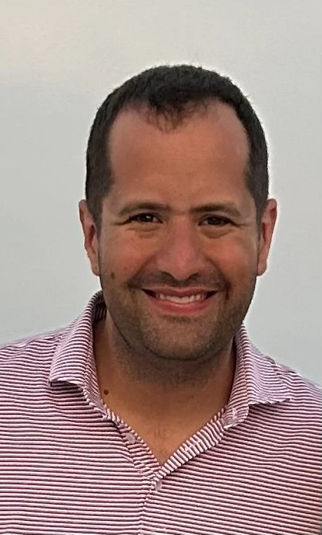A man smiling, wearing a red and white striped collared shirt, against a neutral background.