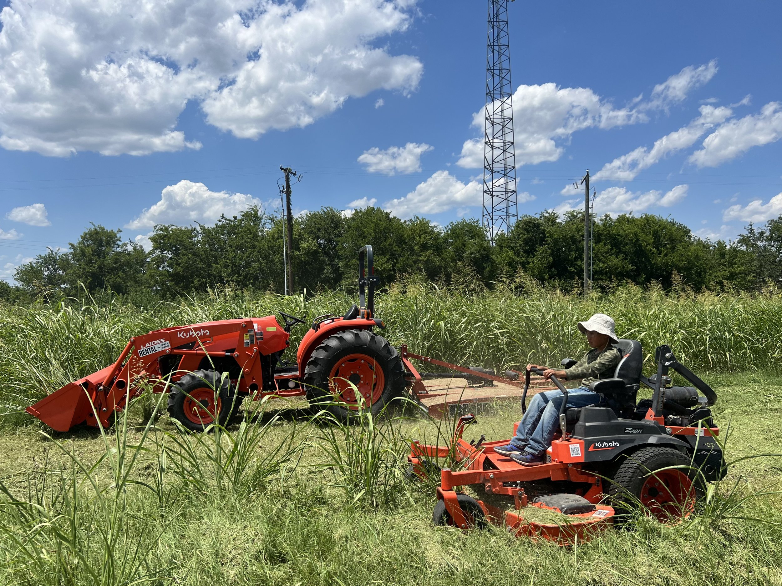 A person operating a Kubota riding mower on a grassy field, with a tractor in the background, under a partly cloudy sky.