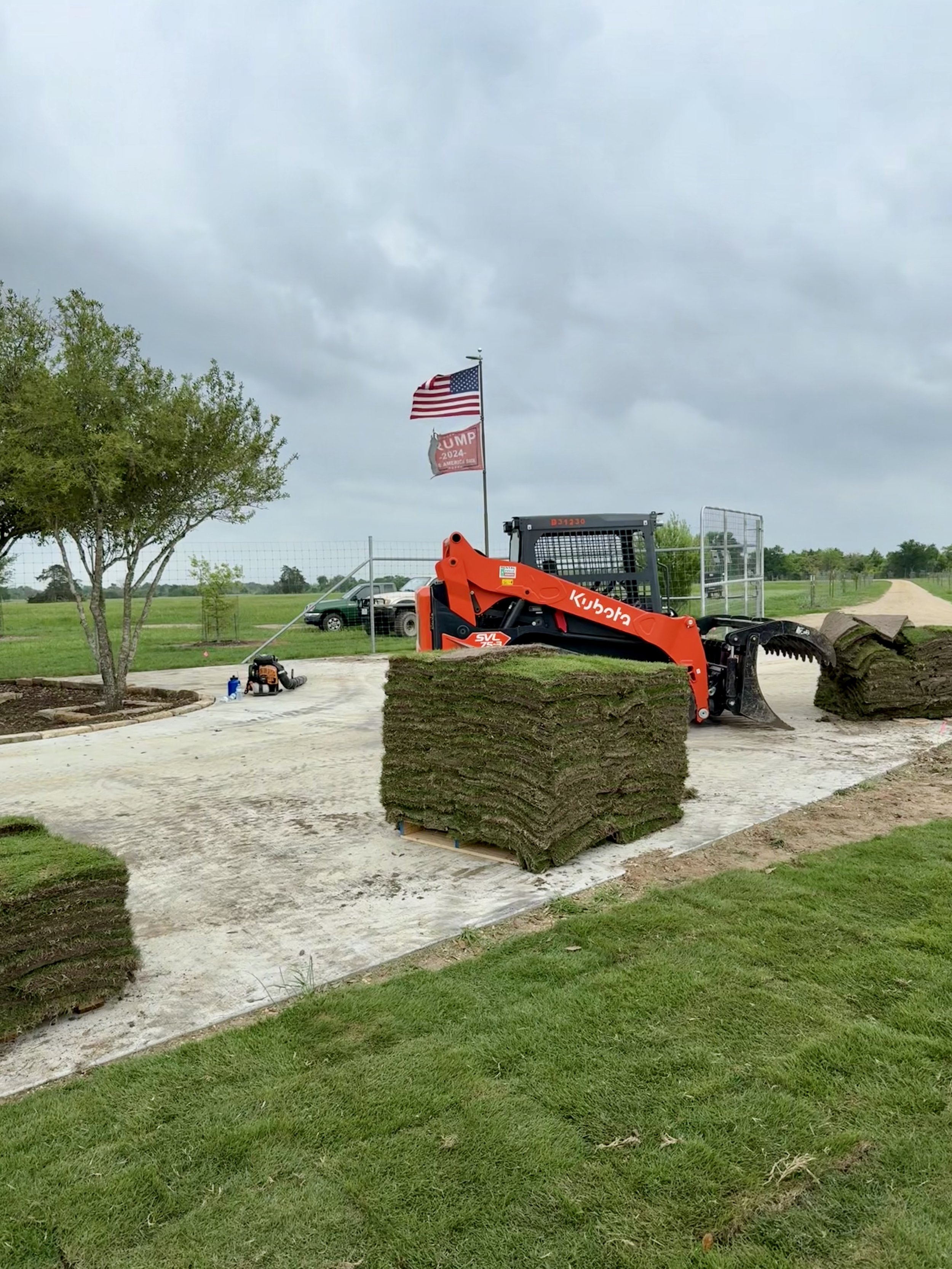 A yard work scene with a Kubota small excavator, a stack of sod rolls on a pallet, a chainsaw and other tools on the ground, and American flags waving in the background. The sky is overcast.