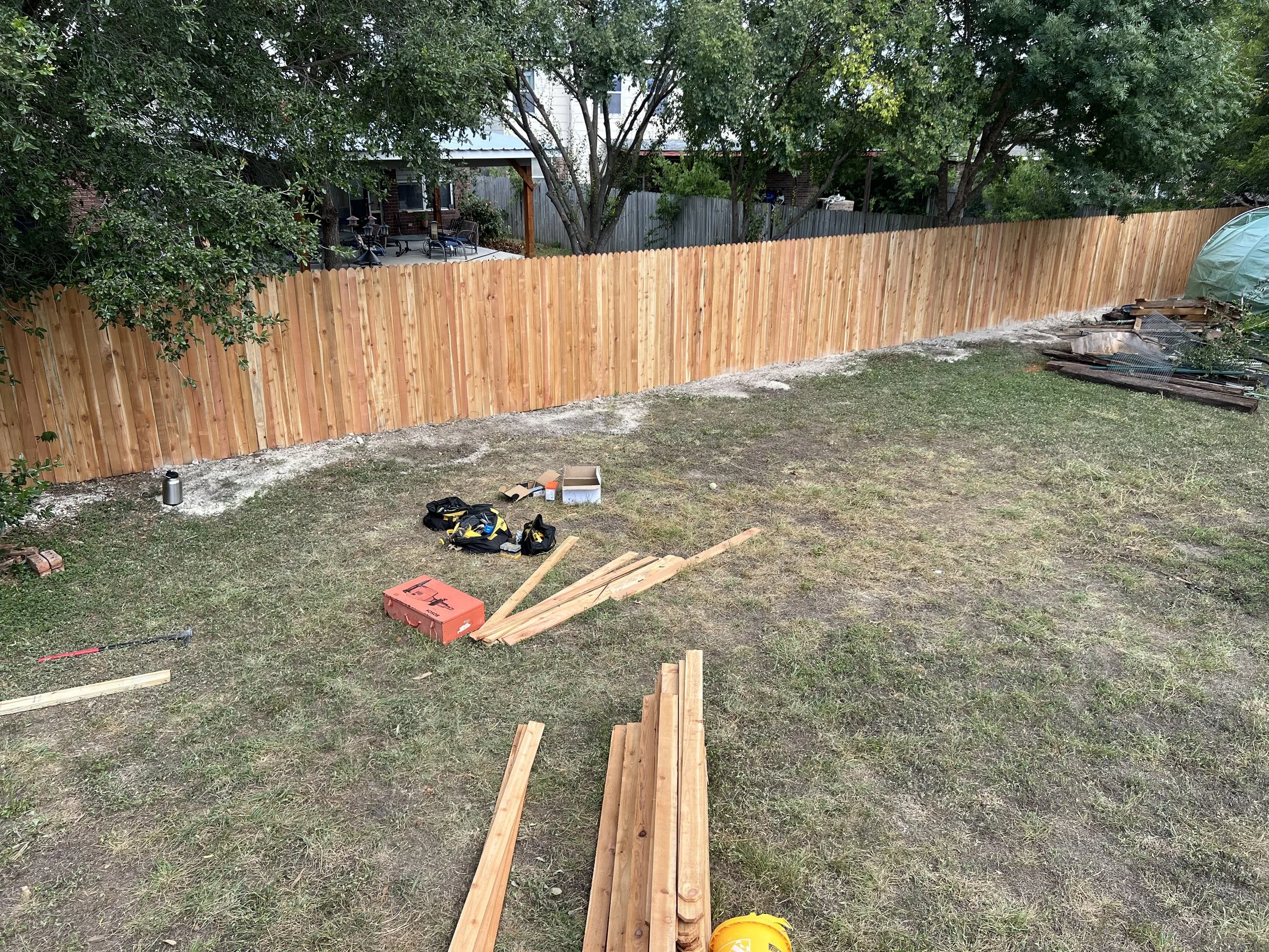 Backyard with a new wooden fence under construction, tools and materials scattered on the grass.