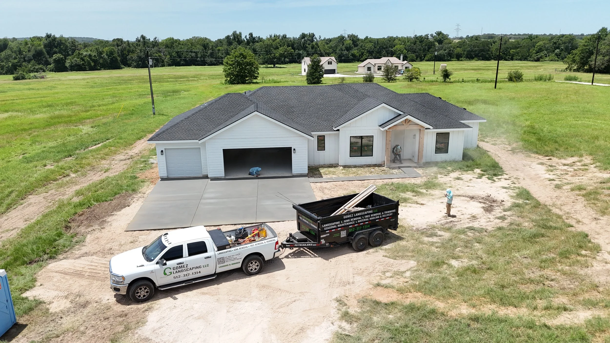 Construction workers are working on a new house with a gray roof and white exterior. A pickup truck with Gomez Landscaping LLC branding is parked in front, hauling construction materials. One worker is kneeling on the driveway, another is near the front door, and a third is standing on the dirt near the house.