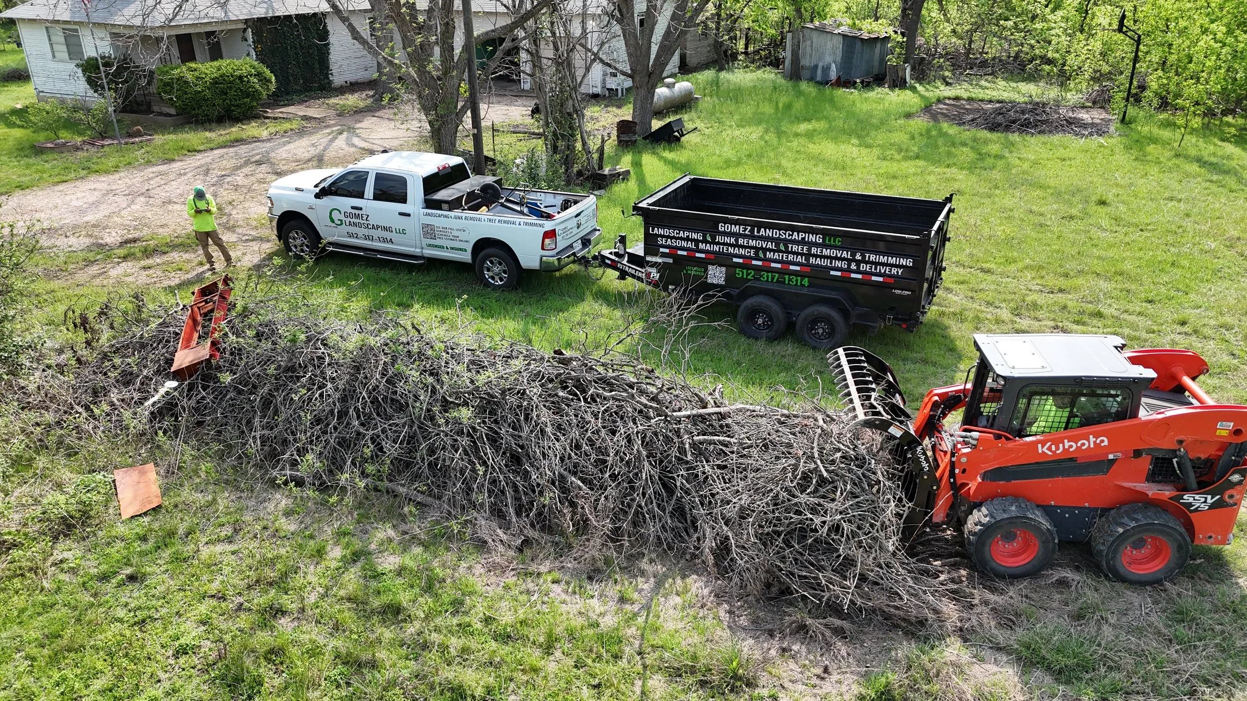 A small orange Kubota skid-steer loader moving a large pile of tree branches in a backyard. Two trucks with company branding are parked nearby, one white pickup truck and one black trailer labeled for landscaping, junk removal, and tree services. A person wearing a green safety vest and hat stands near the trucks, supervising the work. The yard has green grass, trees, and some structures in the background.