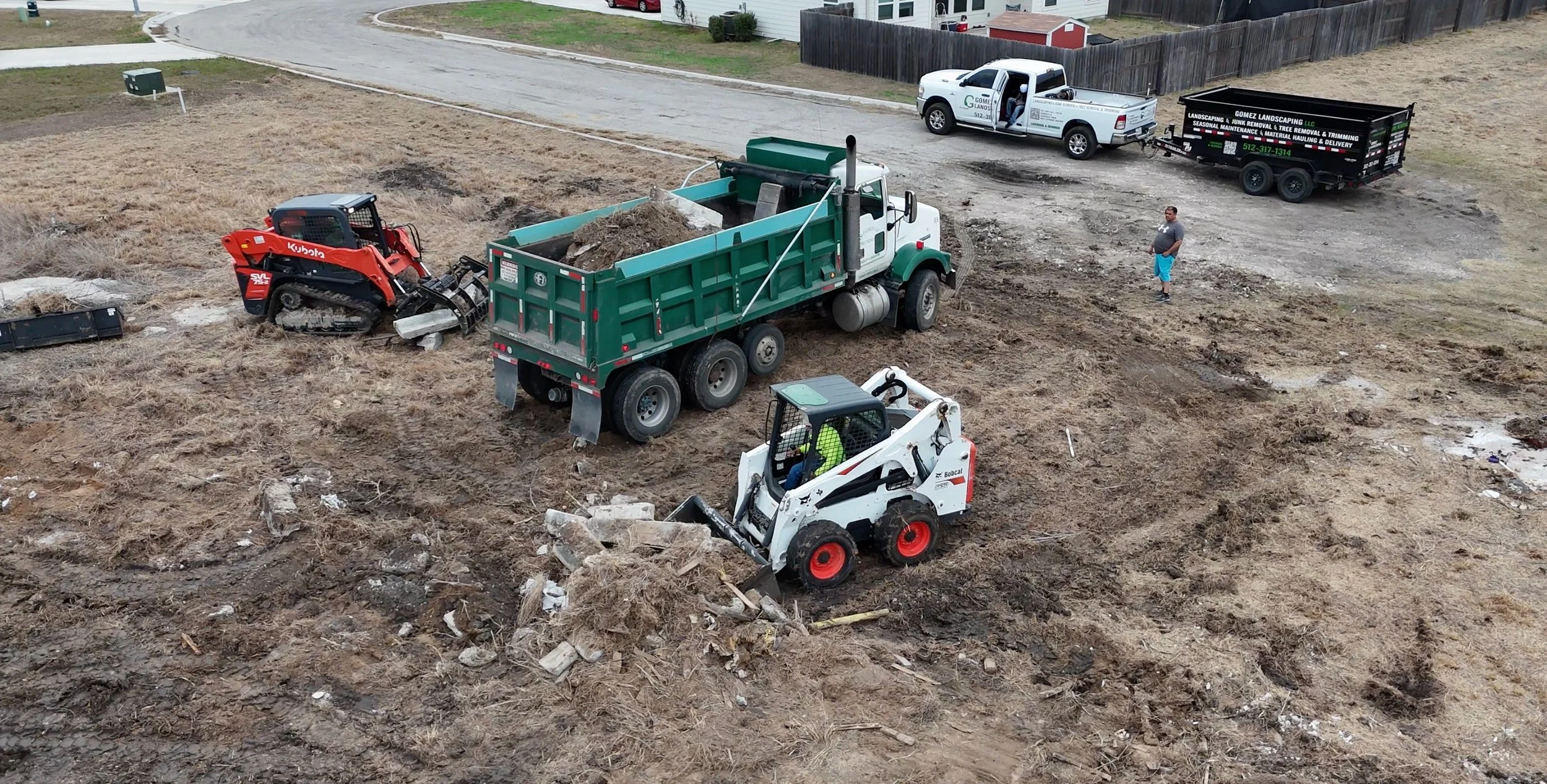 Construction site with machinery and vehicles, including a small Bobcat loader lifting debris, a dump truck, an excavator, and a pickup truck, with a person standing nearby.