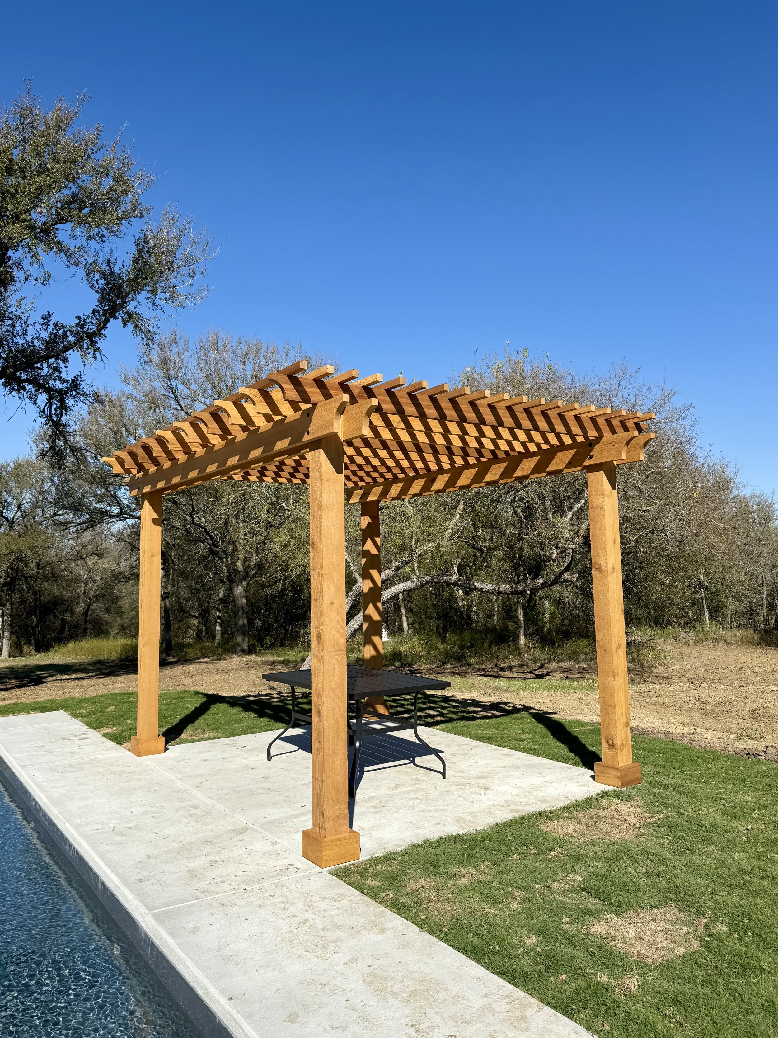 A wooden pergola structure beside a pool, with a table underneath, on a concrete patio, surrounded by grass and trees, under a clear blue sky.
