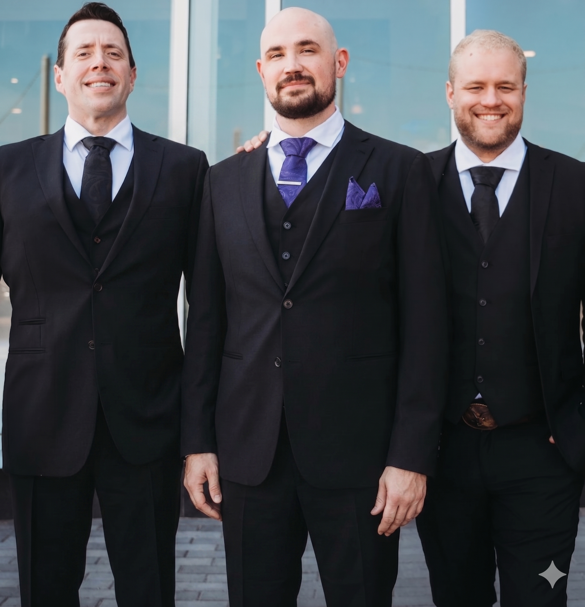 The owners of Frost Leaf, three men, in black suits, white shirts, and black ties, standing outside a modern glass building, posing for a group photo.
