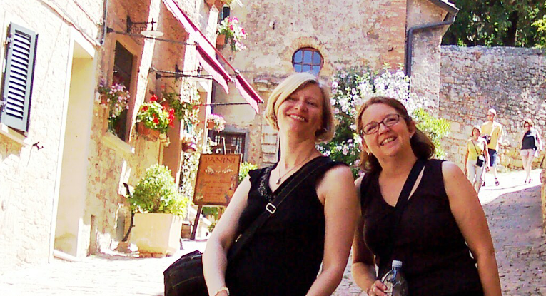 Two women smiling and standing on a cobblestone street in a Mediterranean town, with colorful flowers and historic buildings in the background.