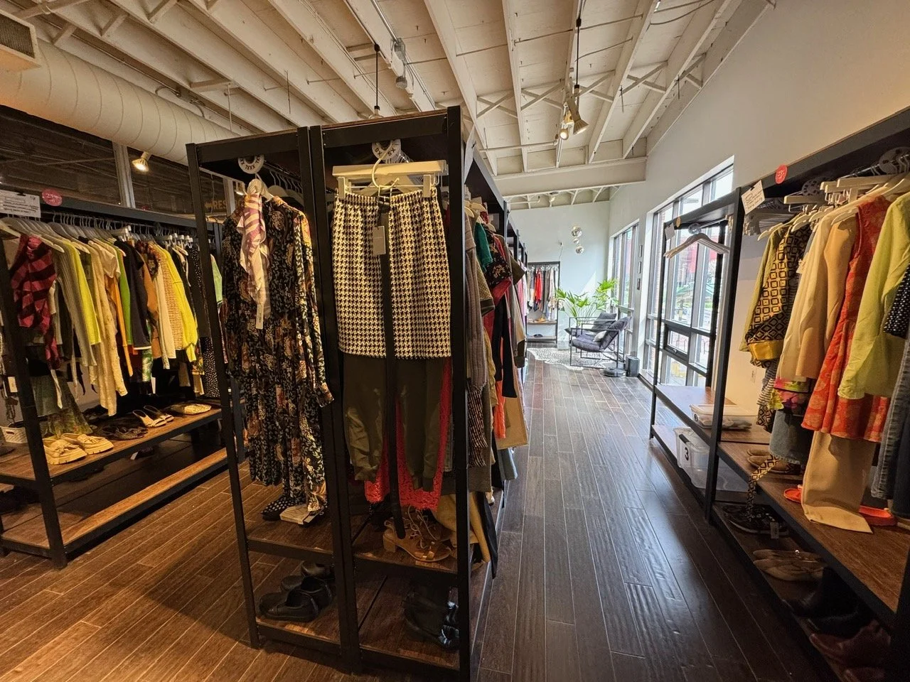 Clothing store interior with racks of clothes, shoes, and accessories, large windows, and seating area in the background.