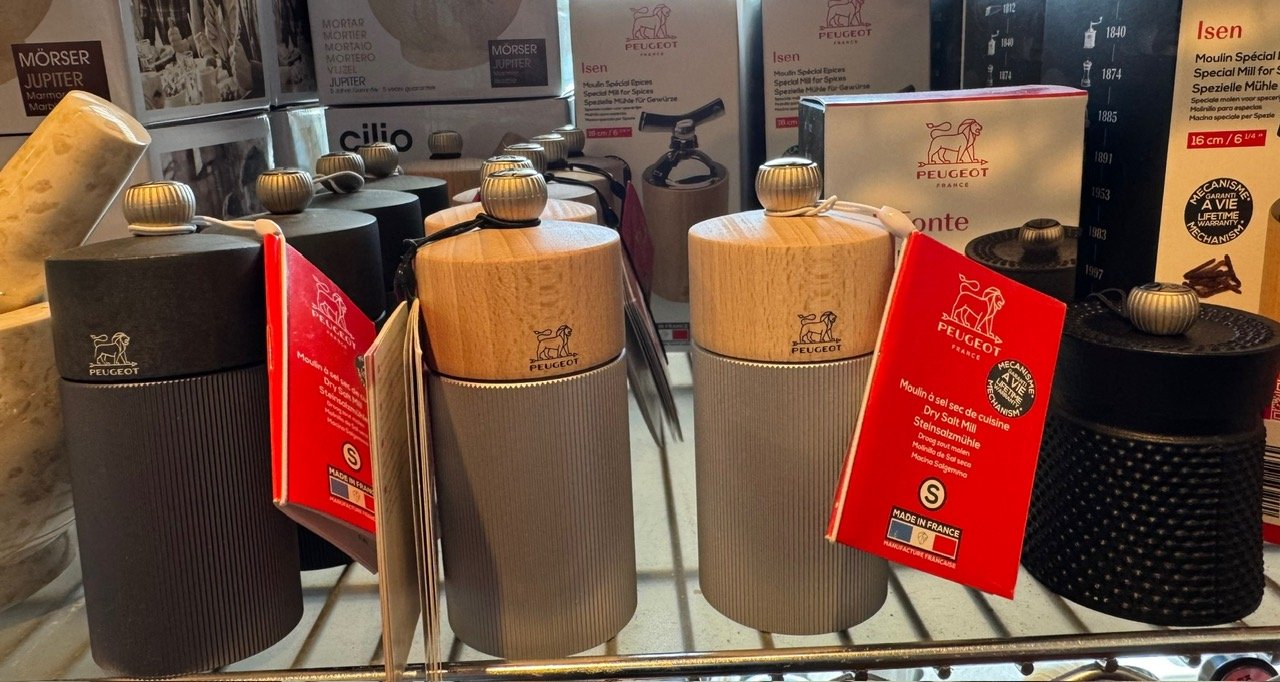 Four Peugeot salt grinders with wooden tops and grey and black bases, displayed on a store shelf, with three on each side of a similar set.