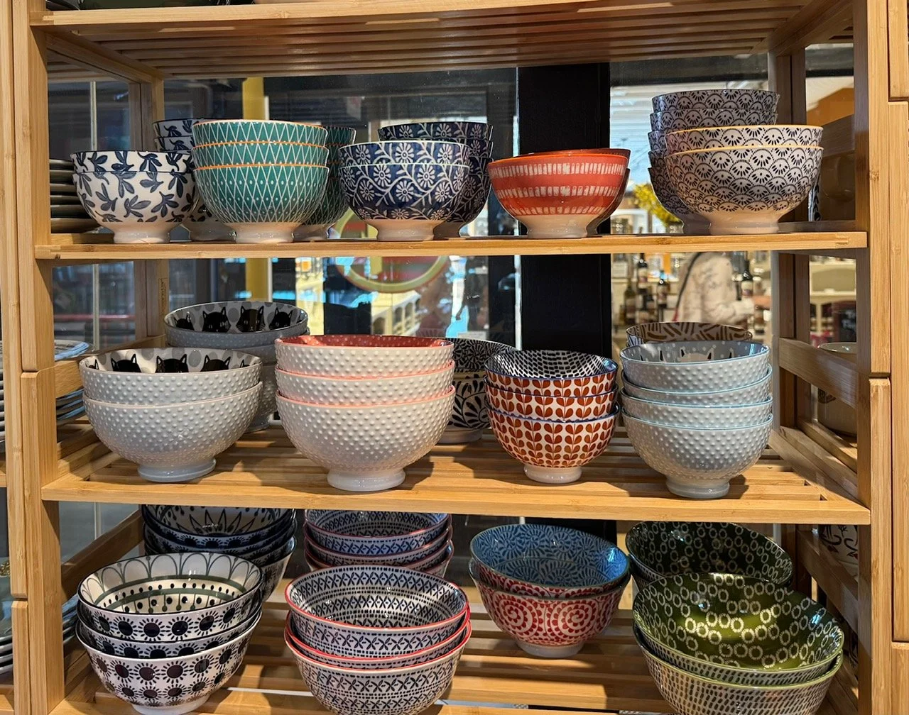 A wooden display shelf holds multiple colorful ceramic bowls with various patterns, arranged in rows inside a store.