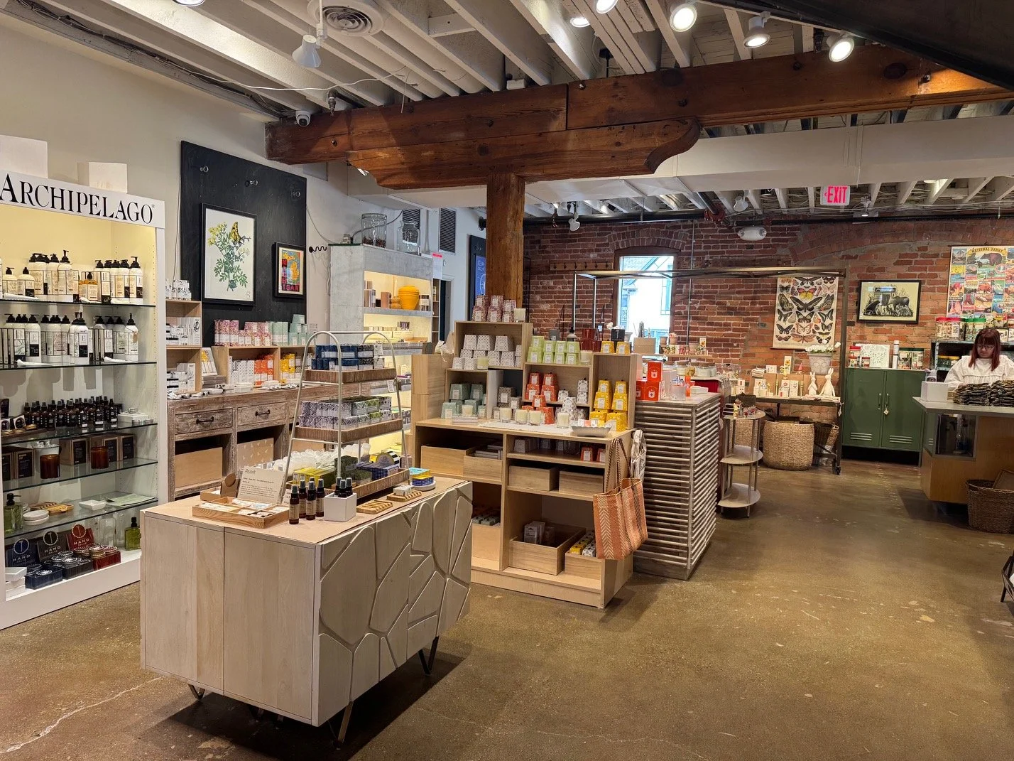 Interior of a store selling personal care and wellness products, with shelves of bottles, soaps, and skincare items arranged on wooden and glass shelves, exposed brick walls, and a woman browsing in the background.