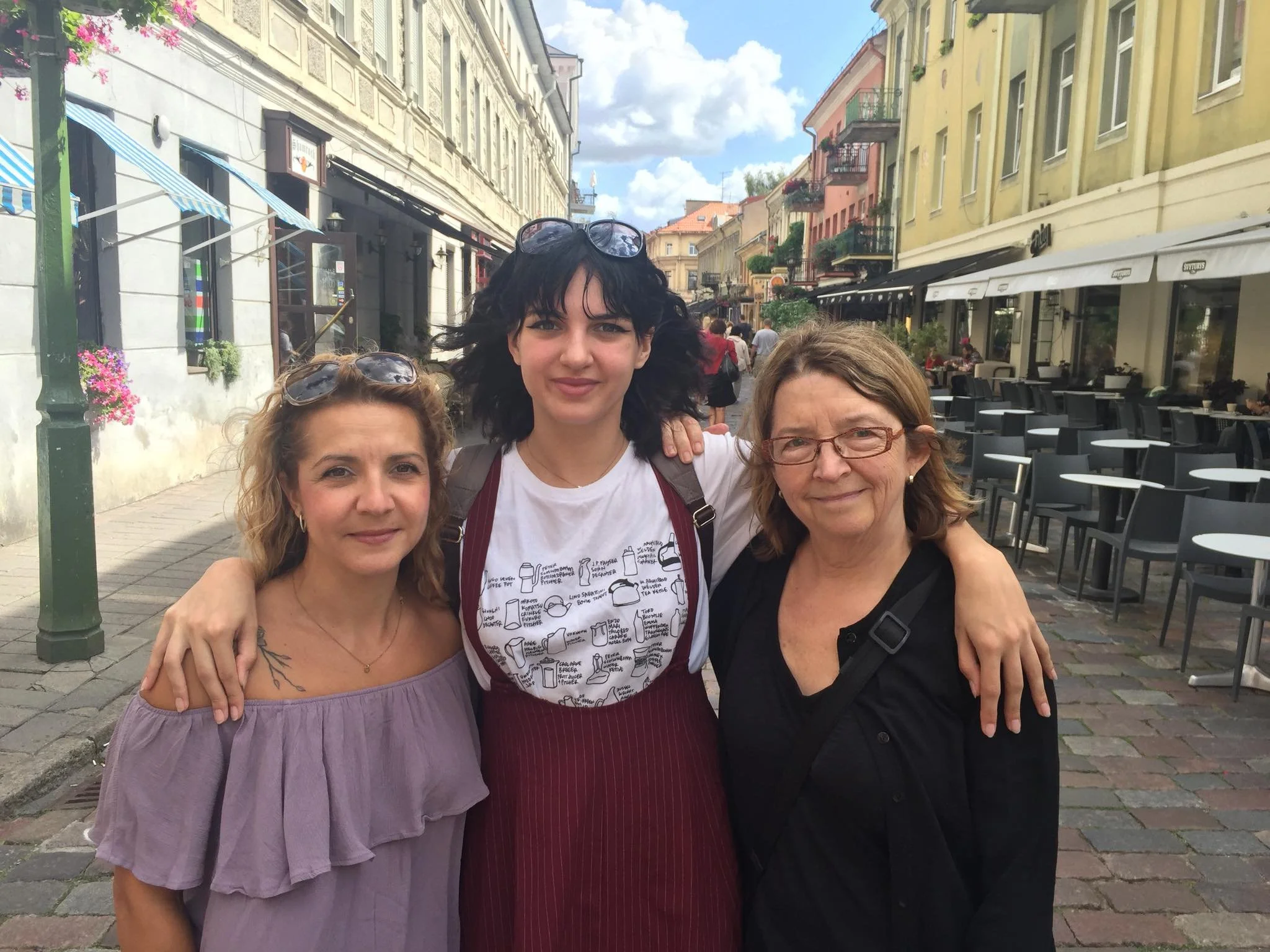 Three women standing close together on a city street, smiling at the camera with colorful buildings and outdoor cafes in the background.