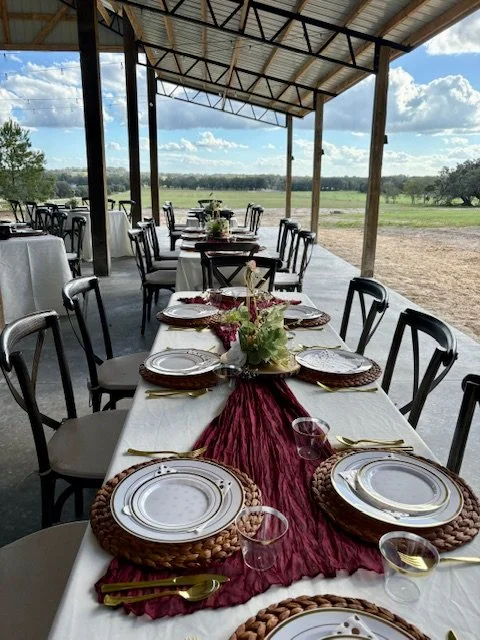 Outdoor event setup with tables under the pavilion. the pavilion. a metal roof, featuring white and burgundy table settings, gold cutlery, and floral centerpieces, overlooking a scenic landscape.