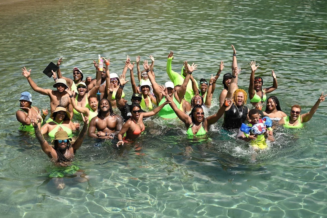 Group of diverse people enjoying a swim in clear water, celebrating with smiles and raised hands on a sunny day.