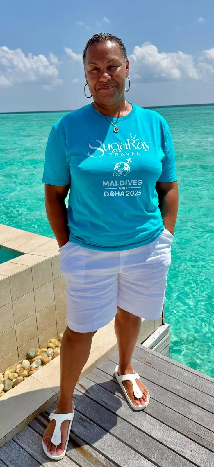 Woman standing on a dock near turquoise water at the Maldives, wearing a blue T-shirt with travel-themed text and white shorts.
