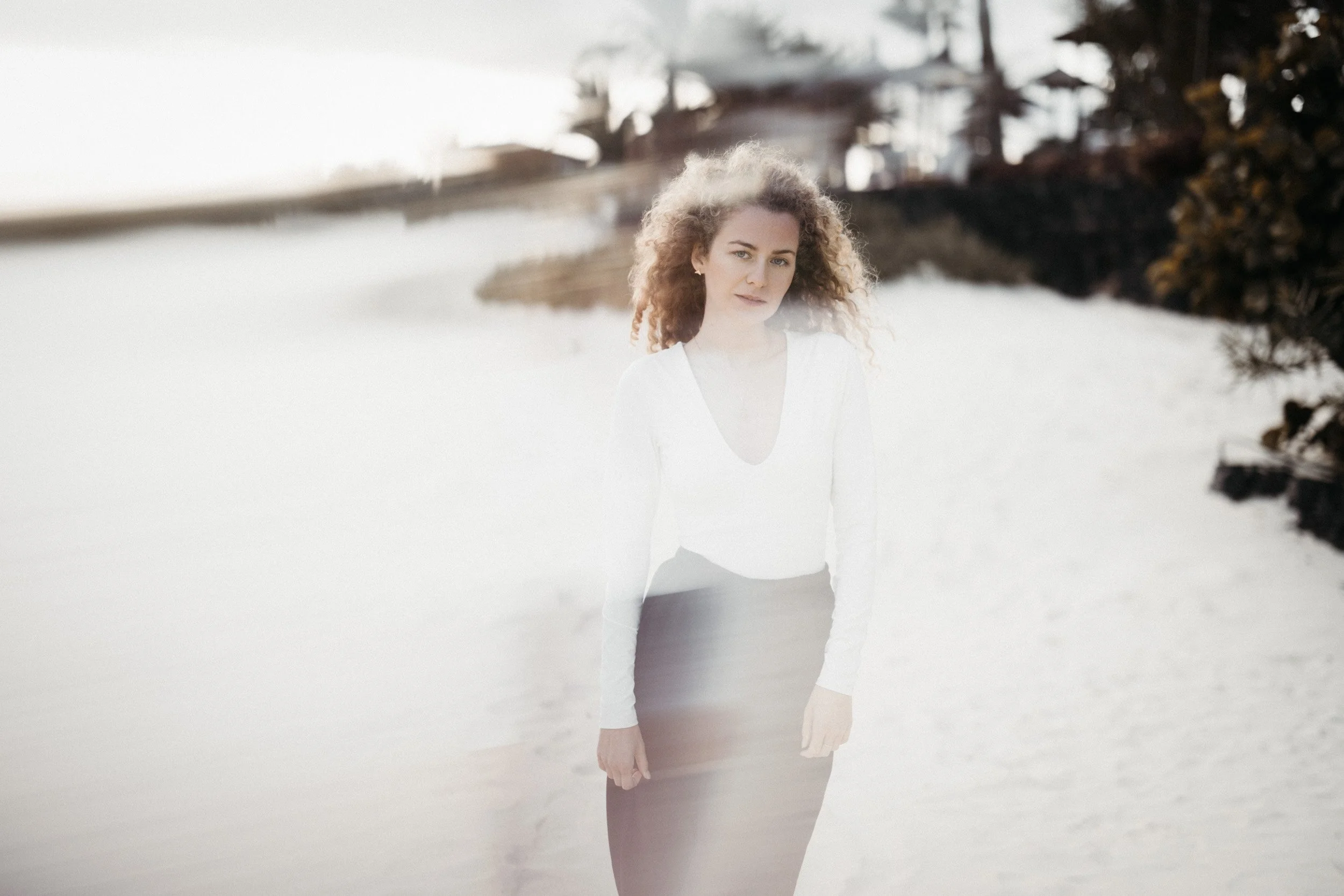 A woman with curly hair wearing a white top and a dark skirt standing on a sandy beach with trees and a house in the background.