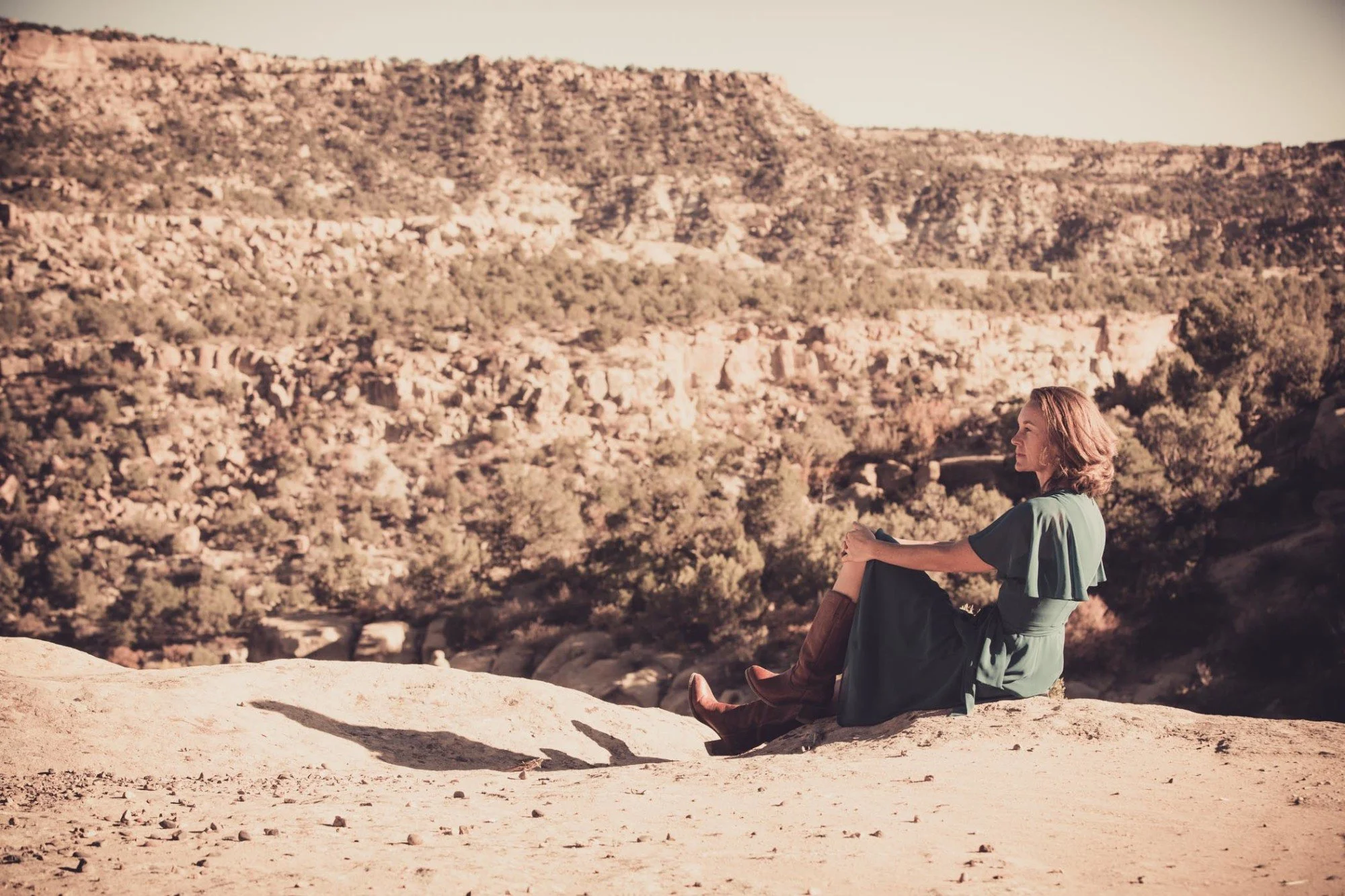 A woman sitting on a rock in a desert landscape, with rocky hills in the background, barefoot, wearing a green dress and brown boots, enjoying a peaceful moment.