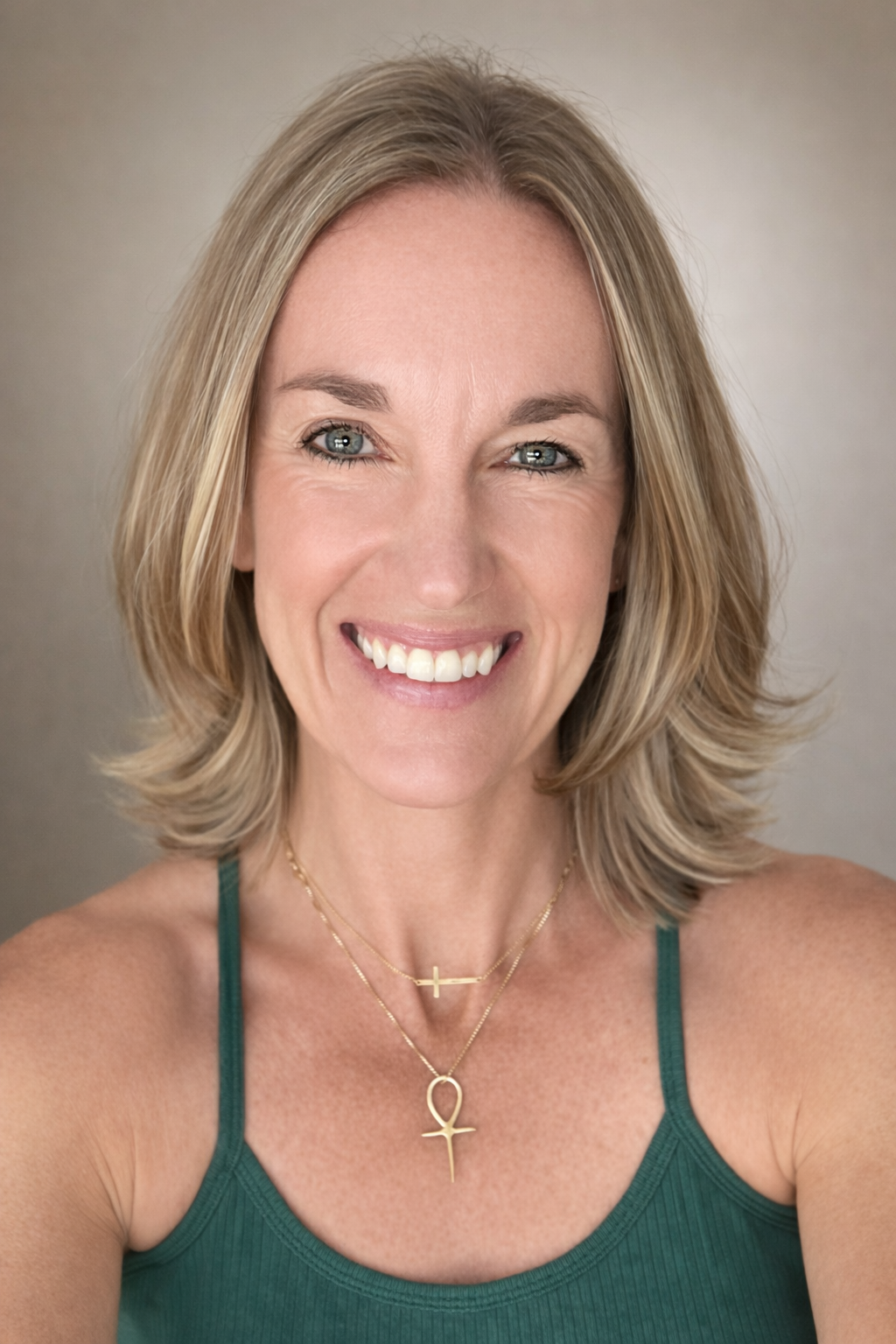 Close-up of a woman with blonde hair smiling, wearing a green tank top and layered gold necklaces with cross pendants.