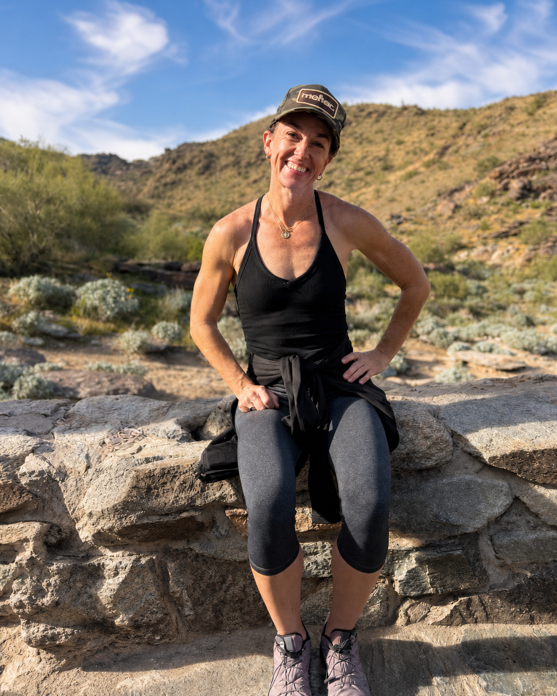 A woman sitting on a rock wall outdoors in a desert landscape with hills and sparse vegetation, smiling at the camera on a sunny day.