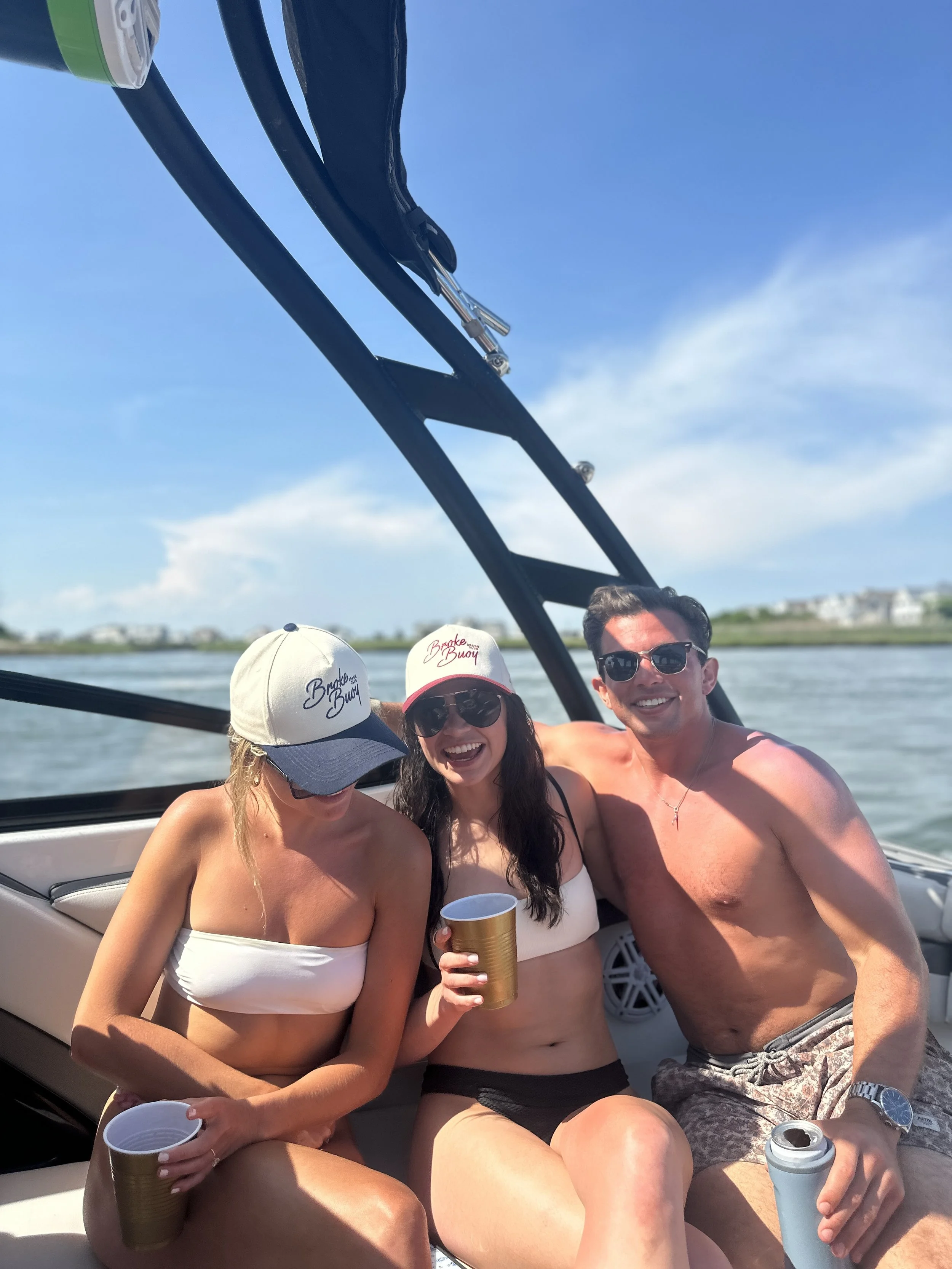 Three friends in swimsuits and sunglasses enjoying a boat ride on a sunny day, holding drinks and smiling.