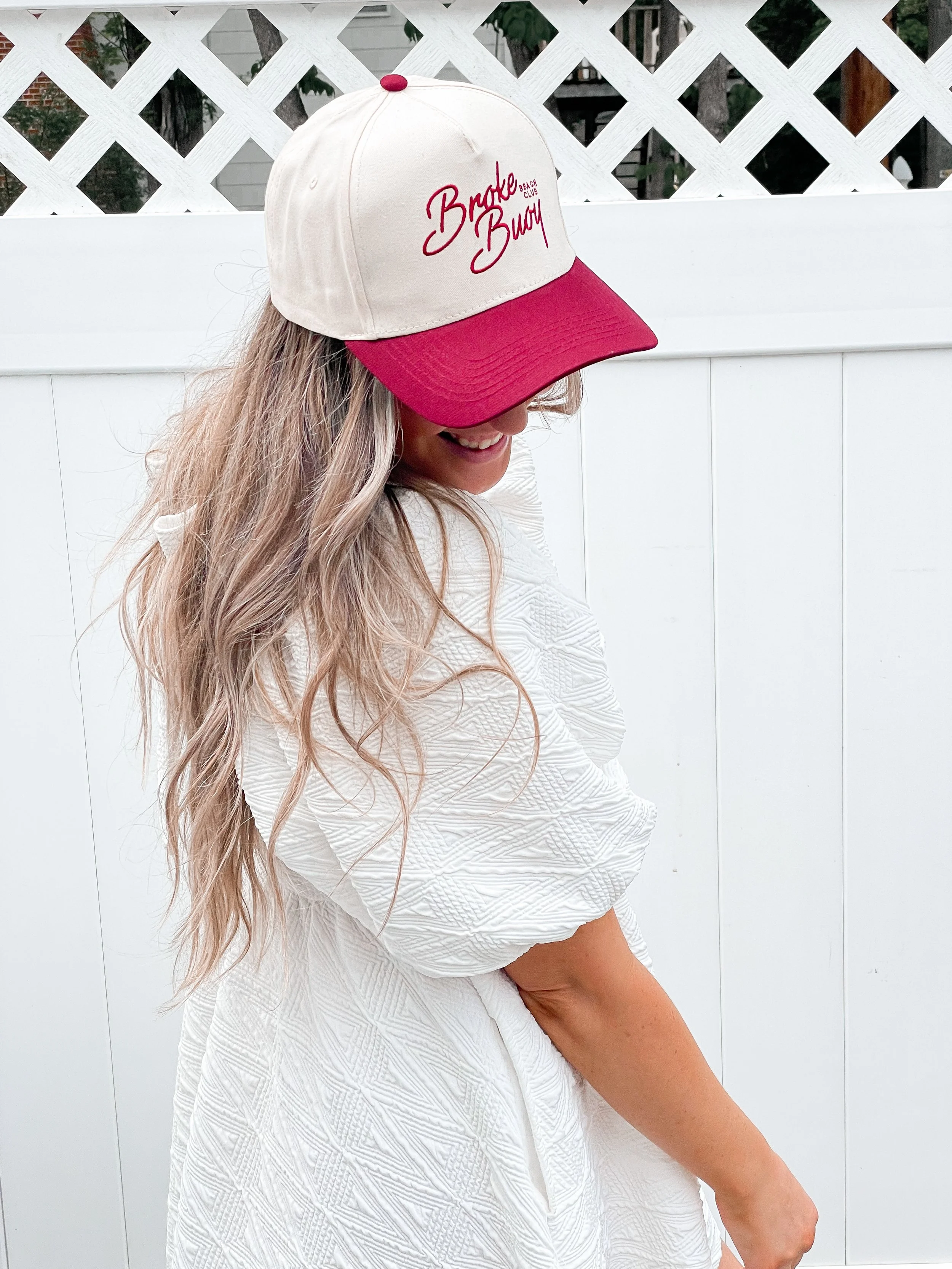 A woman with long, wavy blonde hair wearing a white textured dress and a beige and red baseball cap that reads 'Broke Beach' is sitting beside a white fence, smiling and looking down.