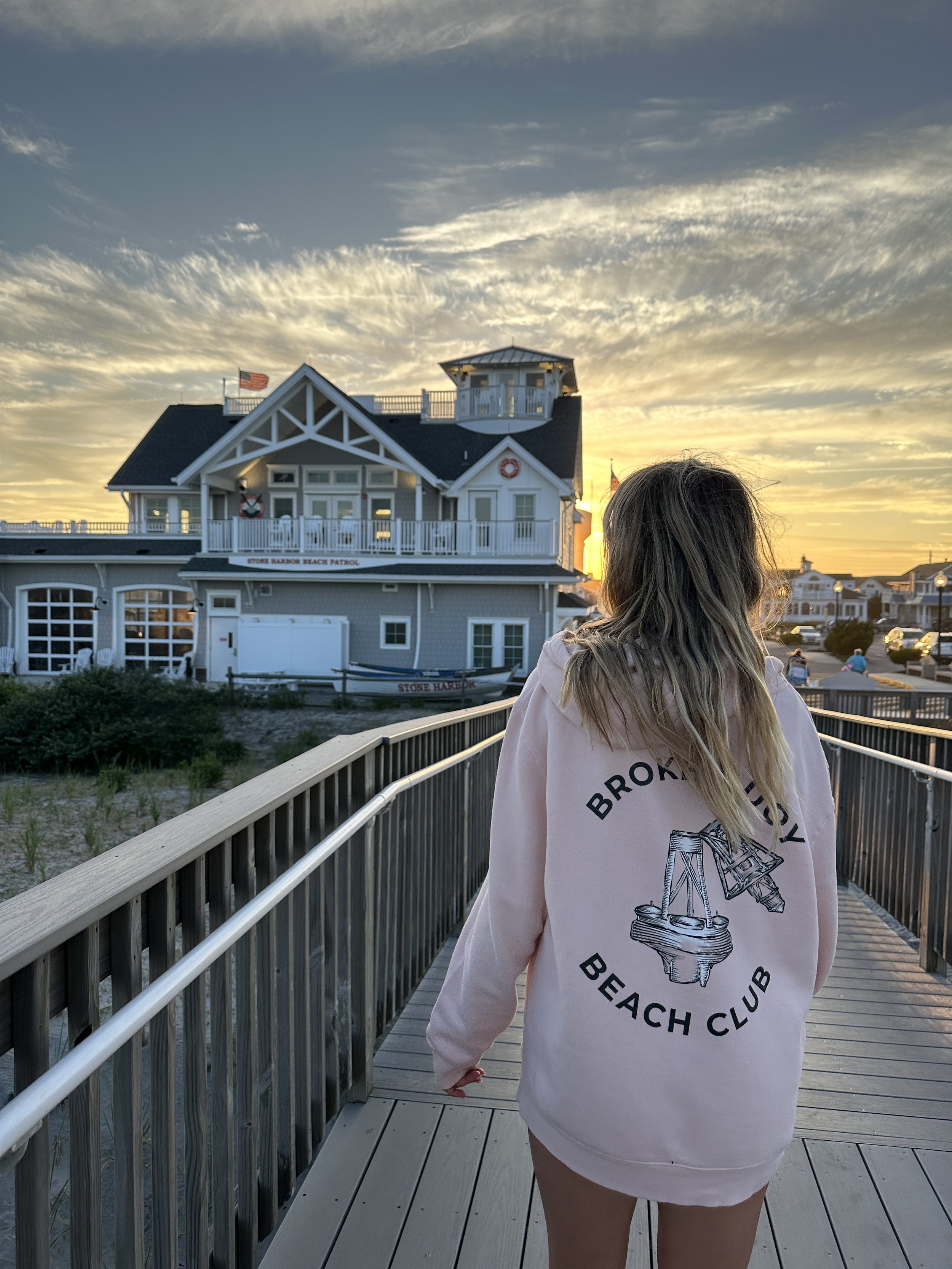 A girl walking on a wooden boardwalk towards a beachfront building at sunset, wearing a pink hoodie with 'Broadway Beach Club' and an illustration of a piano on the back.