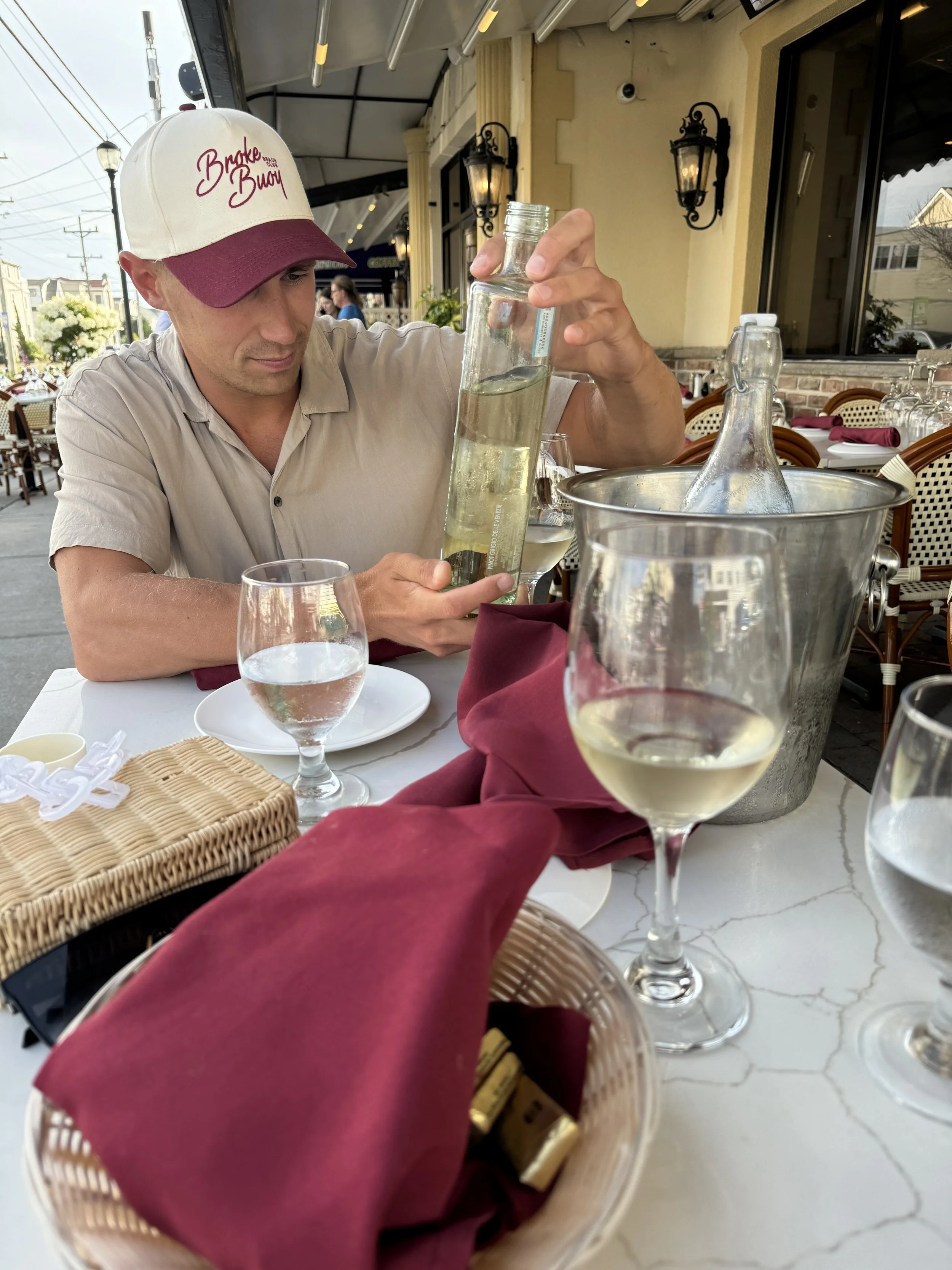 A man wearing a beige shirt and a white baseball cap with maroon accents pouring water into a glass. The setting is an outdoor restaurant with tables, wine glasses, and a wicker basket on the table. The restaurant has a yellow exterior with black dec