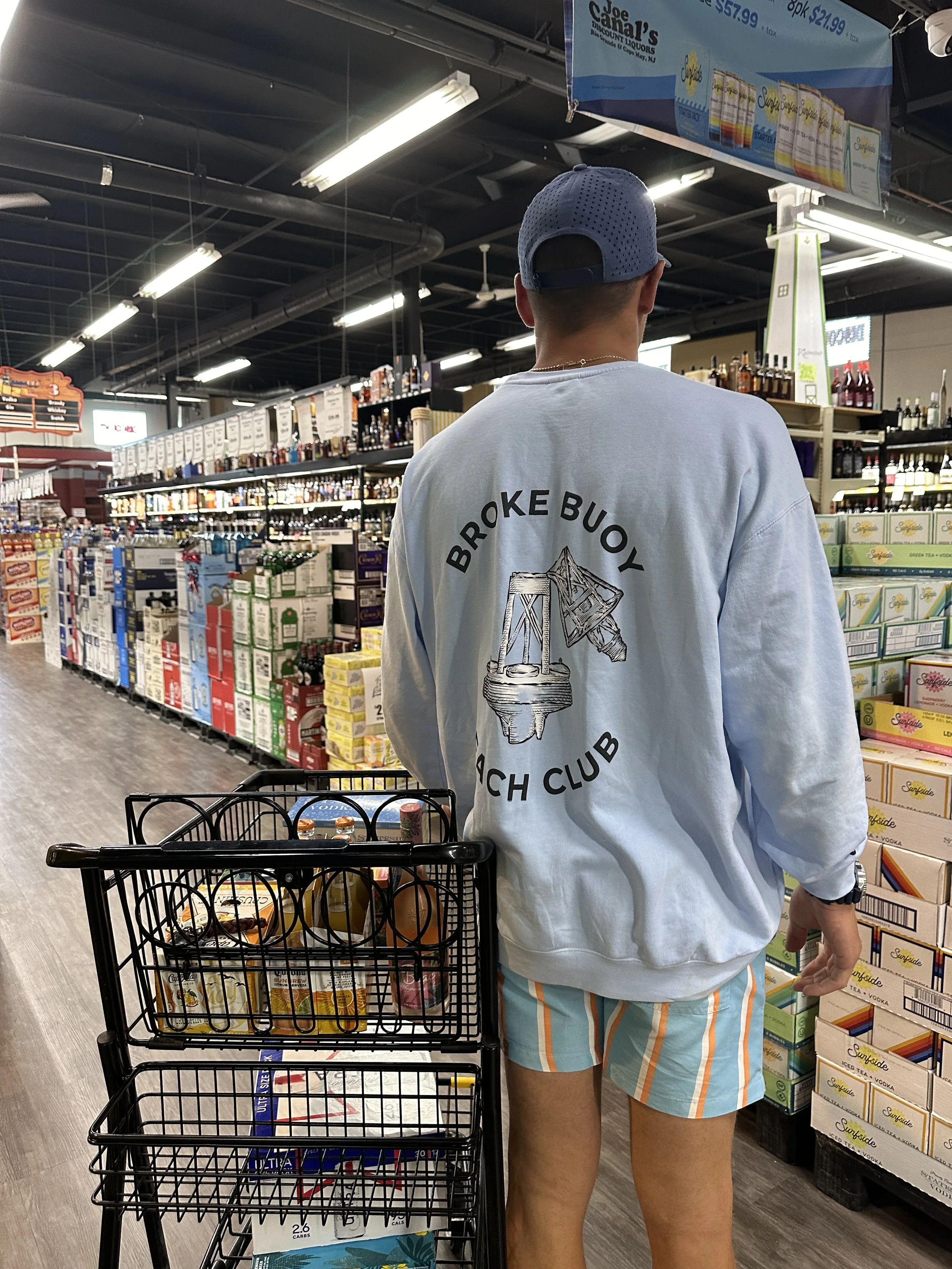 A man in a blue baseball cap, light blue sweatshirt with 'Broke Buoy Beach Club' and a graphic of a buoy, and colorful striped shorts shopping in a grocery store aisle.