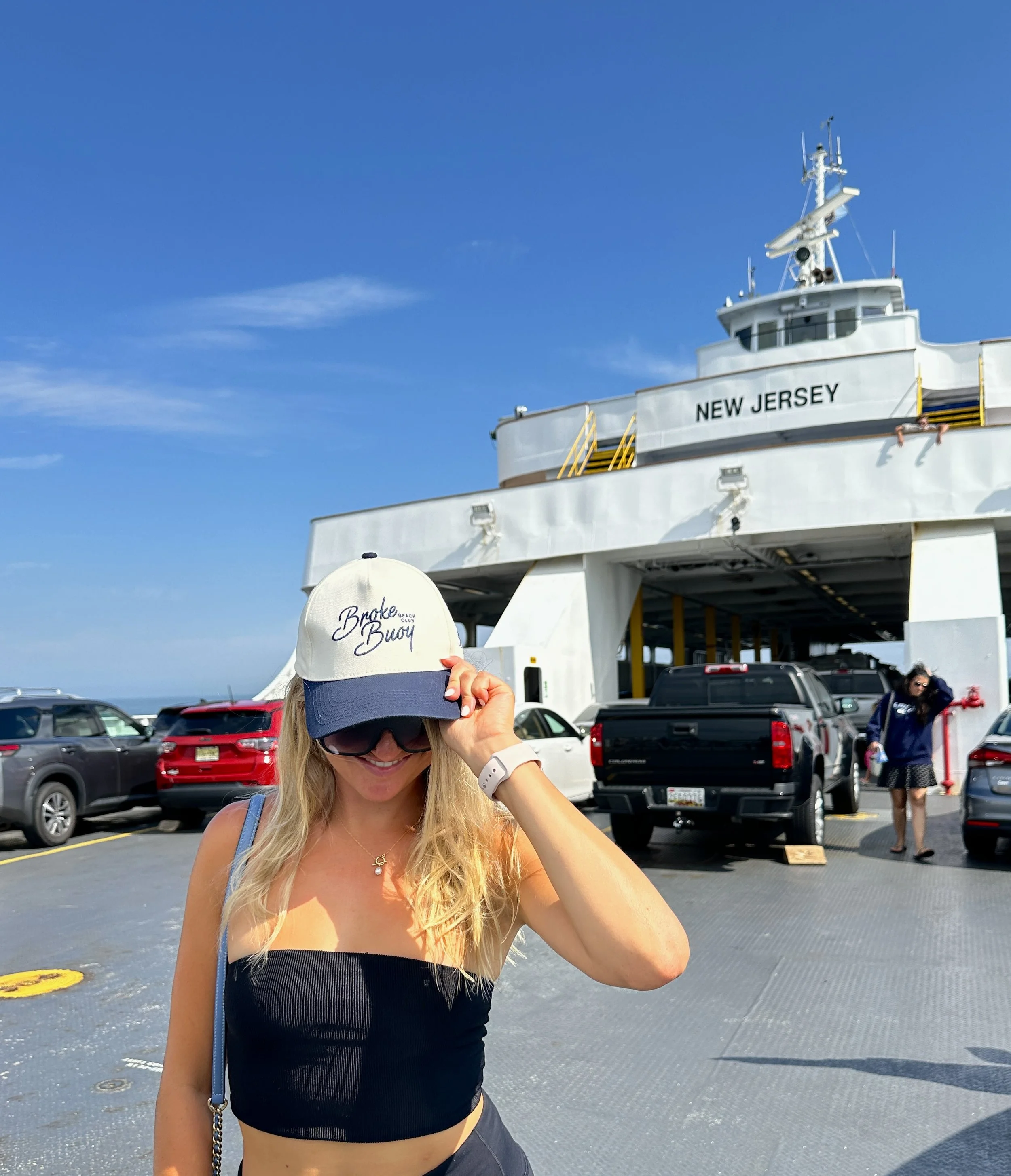 A woman wearing a cream and navy baseball cap with the words 'Broke Buoy' on it, sunglasses, and a black tube top, standing in front of a ferry named 'New Jersey,' with parked cars and another woman in the background, under a blue sky.