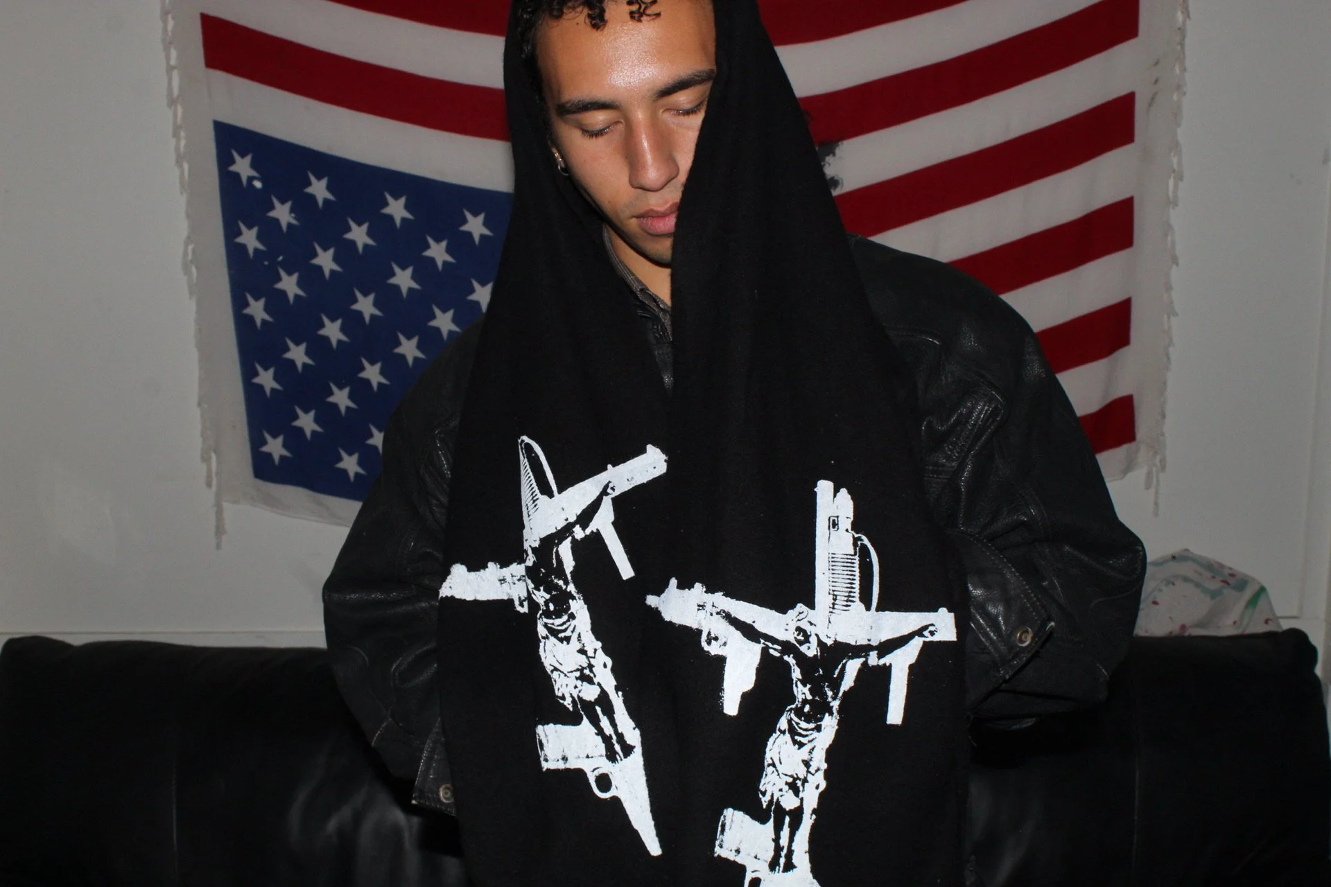 A young man wearing a black hoodie with white guns graphic, standing in front of an American flag hanging on the wall.