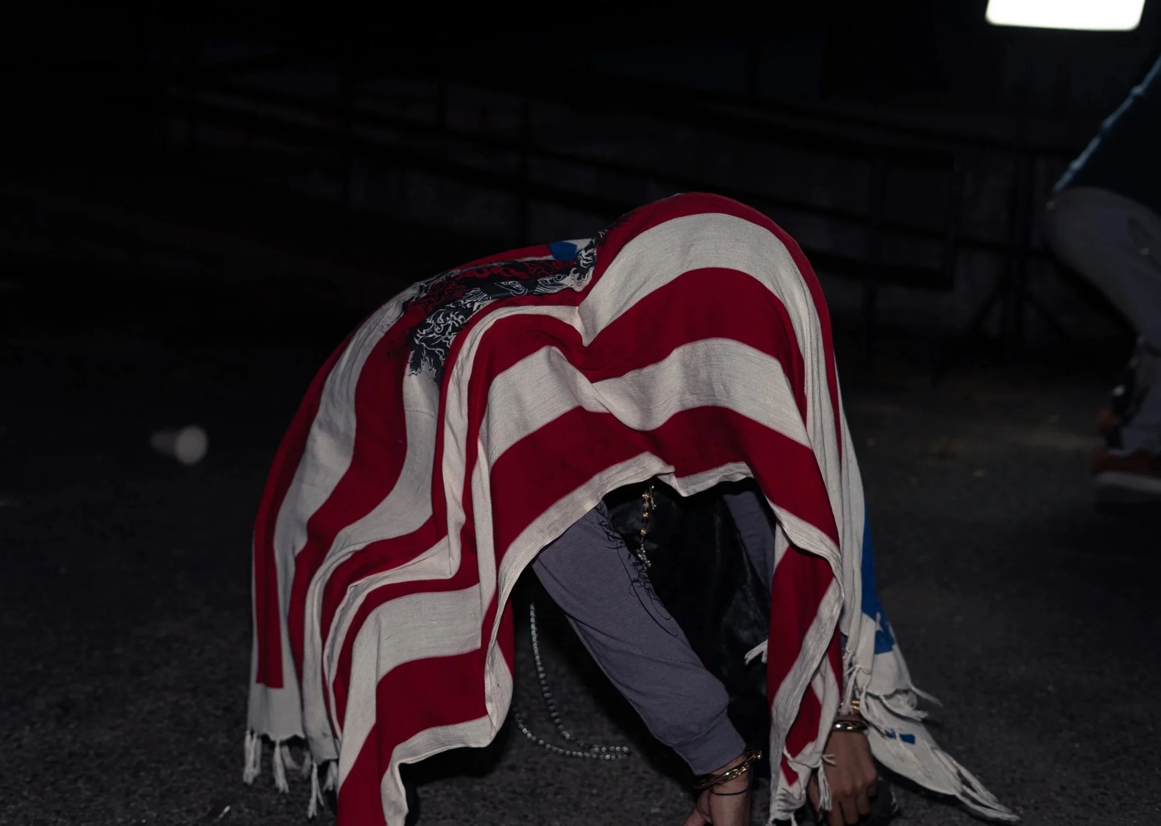 Person crouching on the ground partially covered with a large American flag, with dark background and minimal lighting.