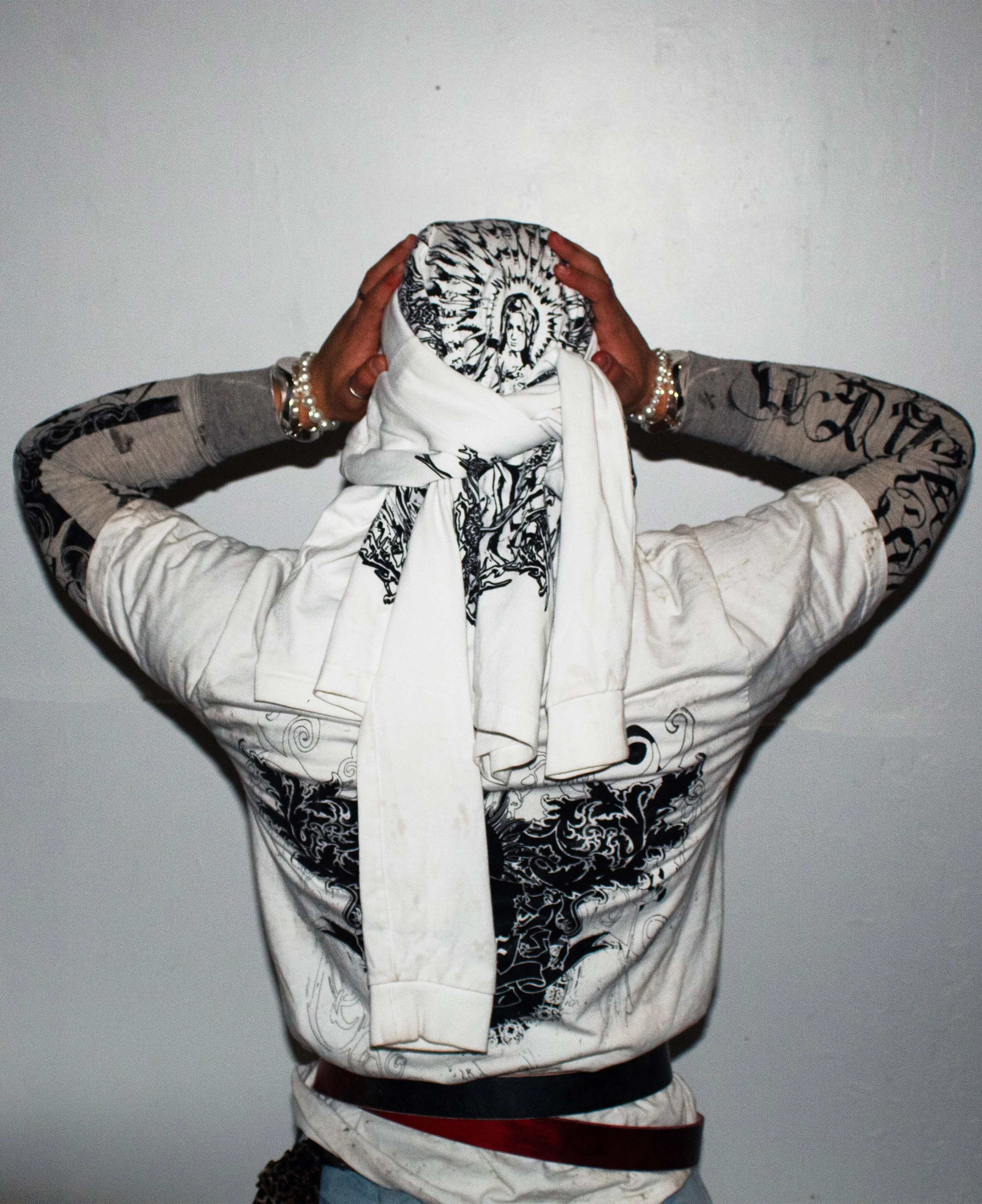 Person with arms raised, wearing a patterned jacket, a head covering with religious imagery, and jewelry, standing against a plain wall.