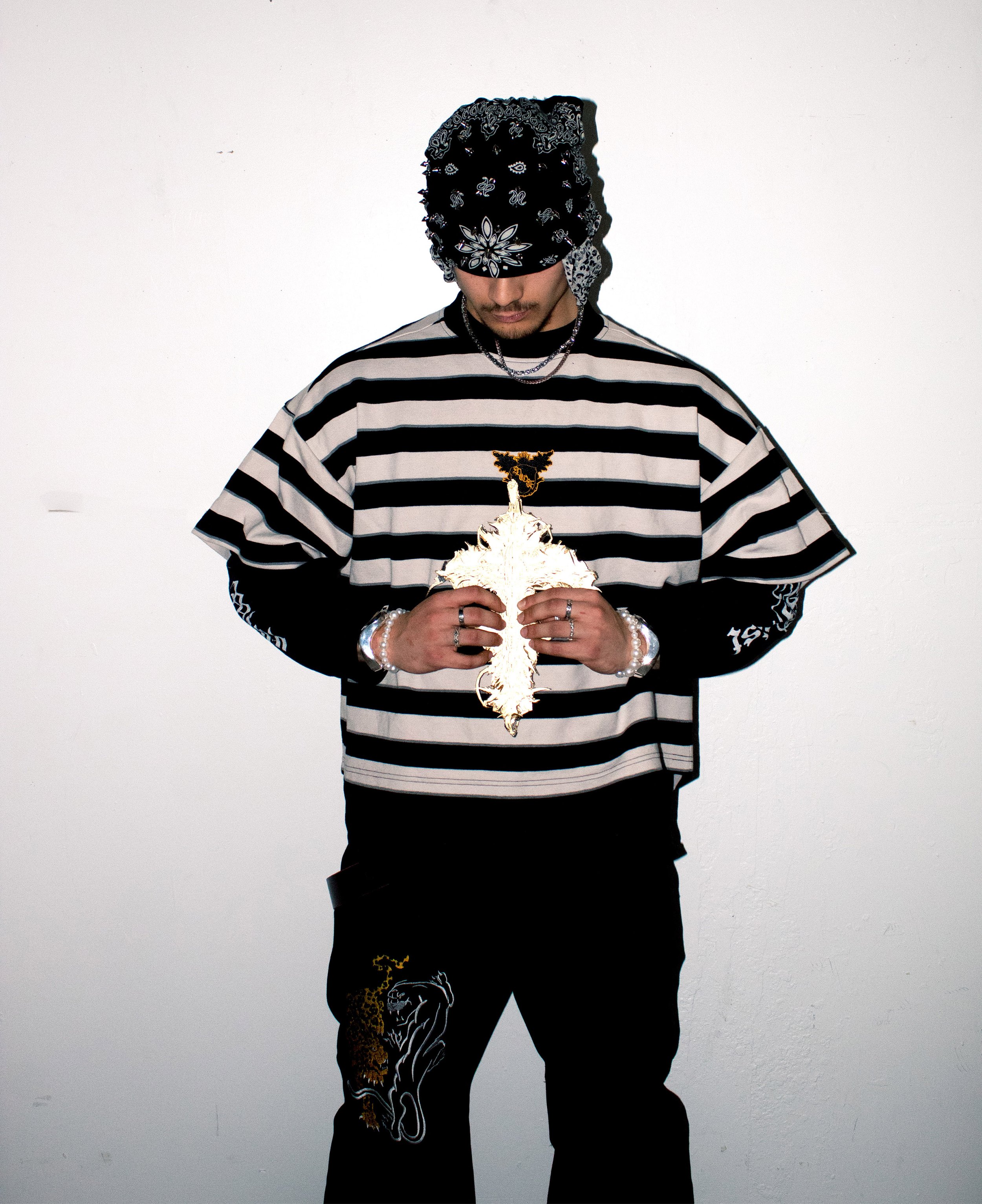 A man wearing a black and white striped shirt, black pants with a lion design, and accessories including rings, bracelets, and layered necklaces. He has a black bandana over his face and is holding a glowing decorative object against a plain white background.