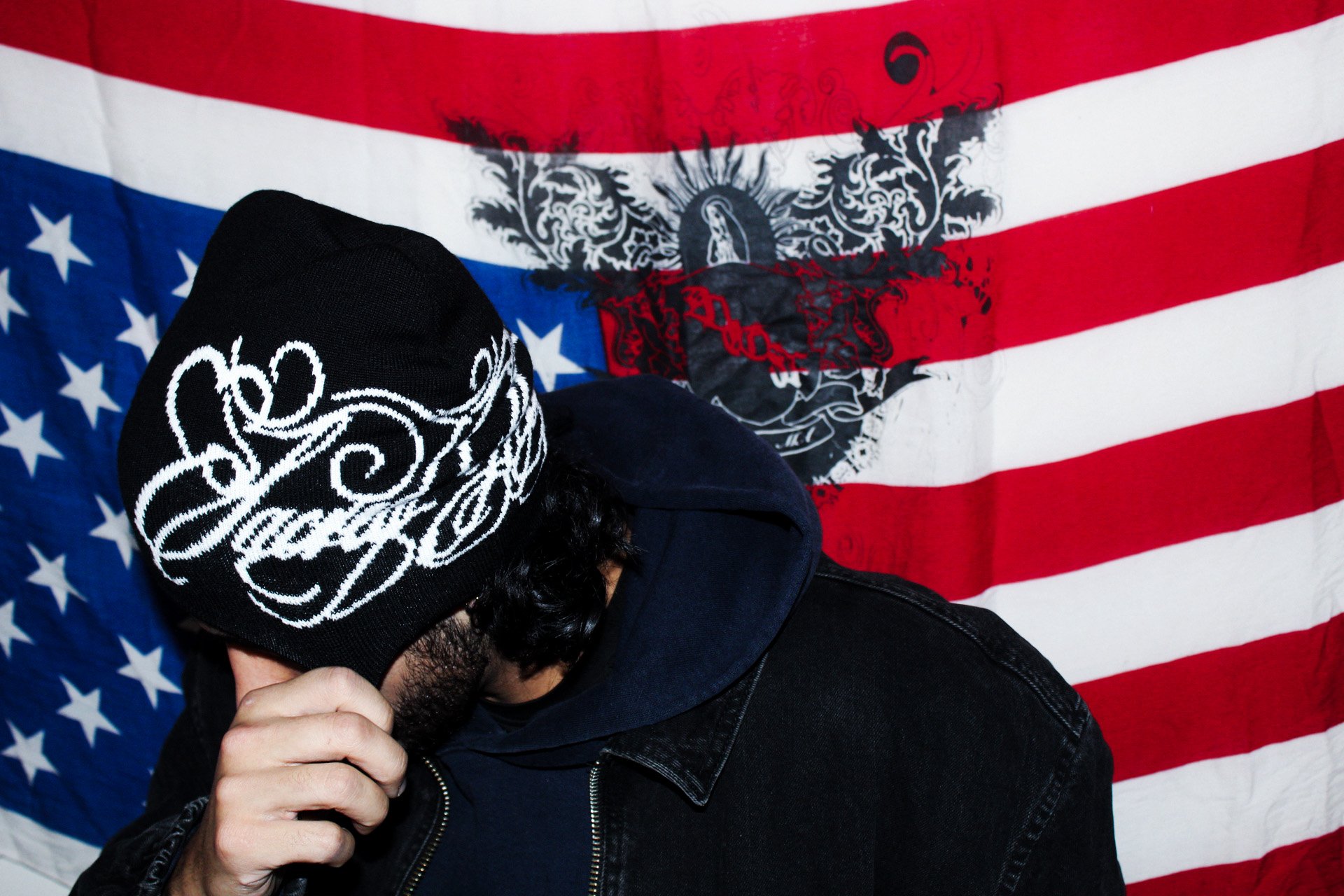 A person wearing a black hoodie and a black beanie with white designs, standing in front of a backdrop of an American flag with a black emblem. The person is touching their face with their hand, looking downward.