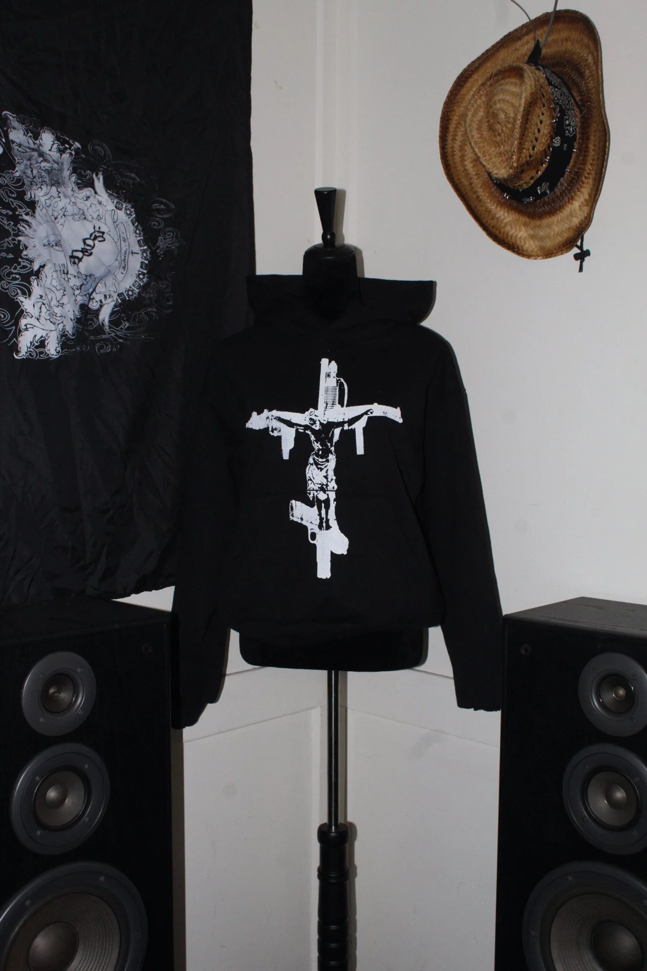 Black hoodie with a white graphic of a crucifix made of guns, displayed on a mannequin. Two large black speakers are on either side of the hoodie. A wide-brimmed straw hat hangs on the white wall above and to the right of the hoodie, and black fabric with a white design is partially visible on the left side of the image.