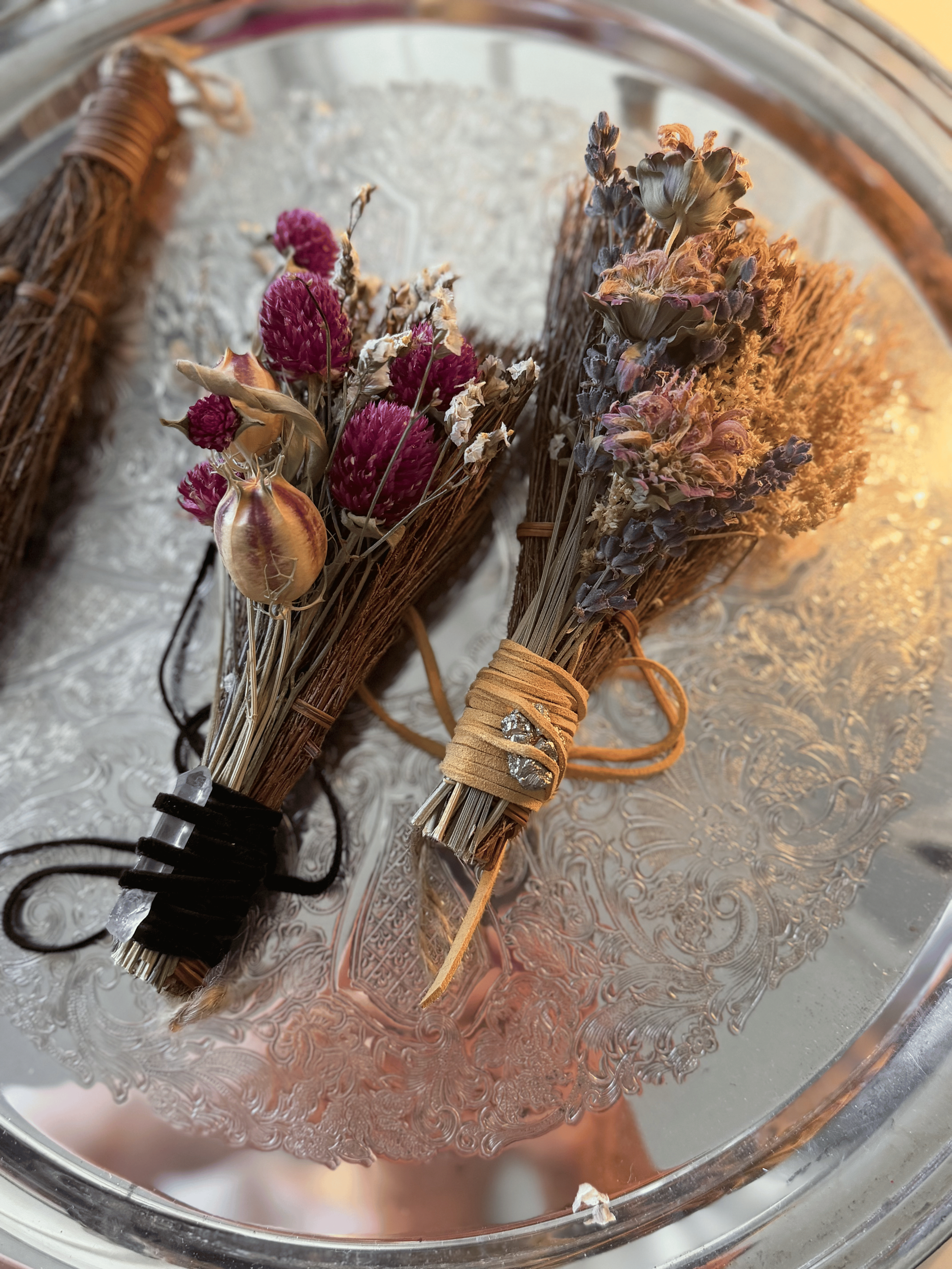 two mini brooms made of cinnamon twigs are adorned with dried florals and herbs and are wrapped with hemp or twine with a raw crystal attached.