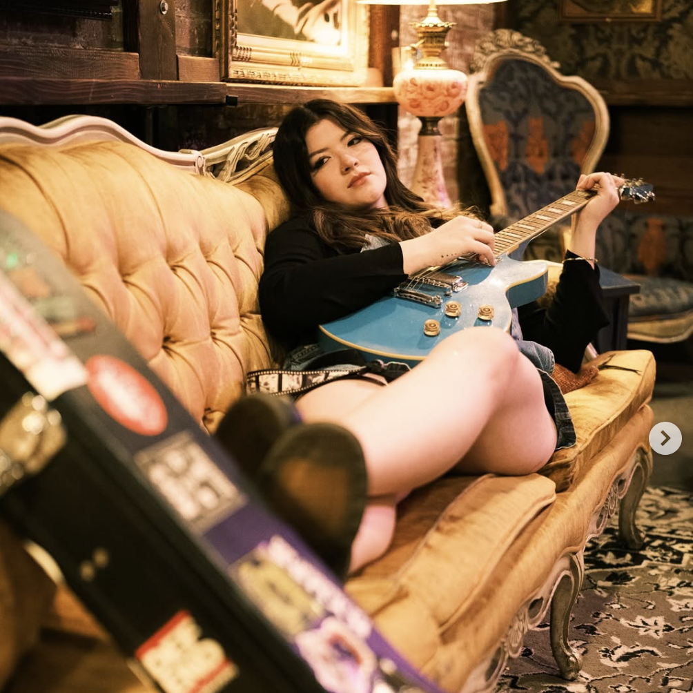 Photo of Hayworth playing guitar while lounging on couch