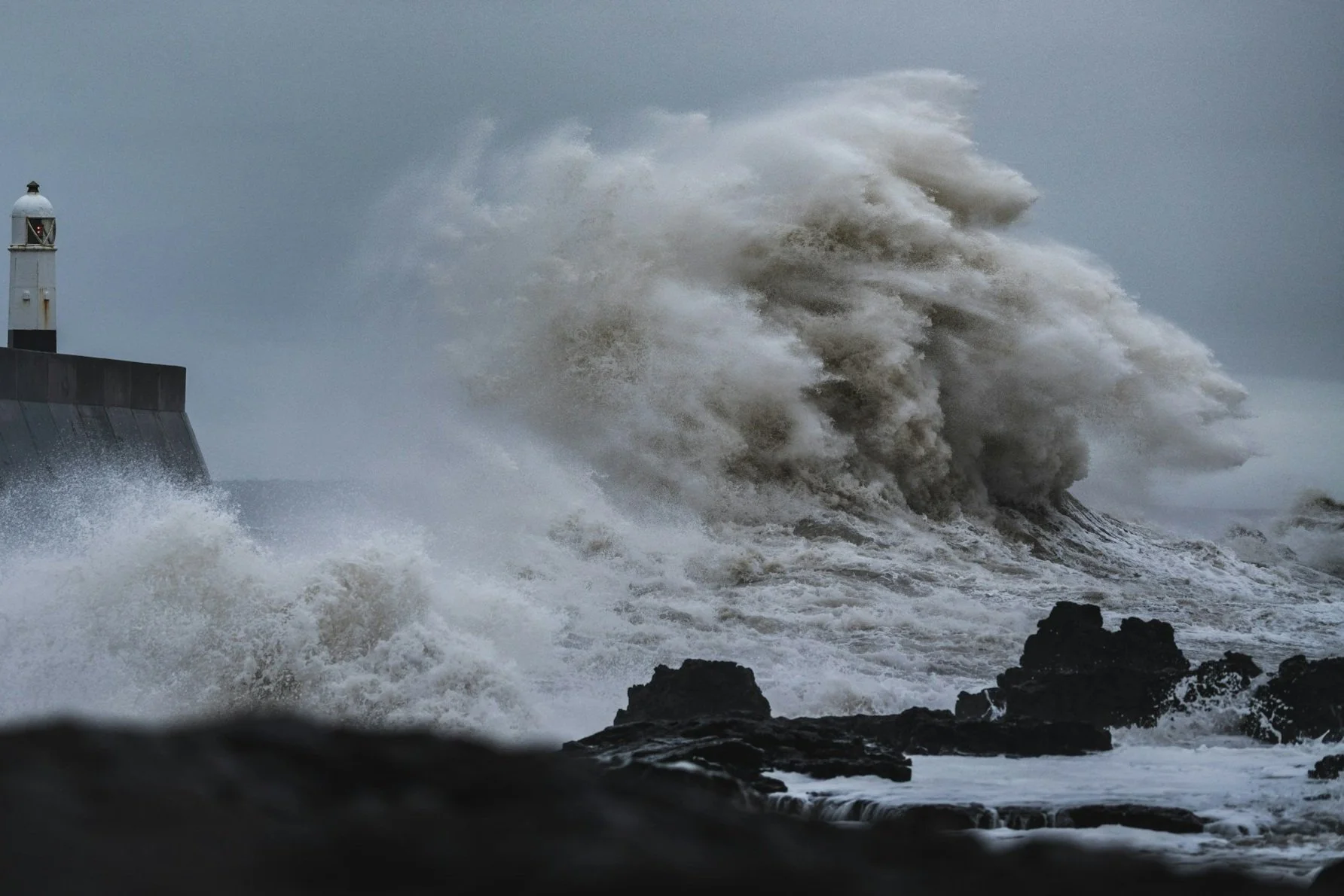 Large waves crashing against a rocky shoreline near a lighthouse on a stormy day.